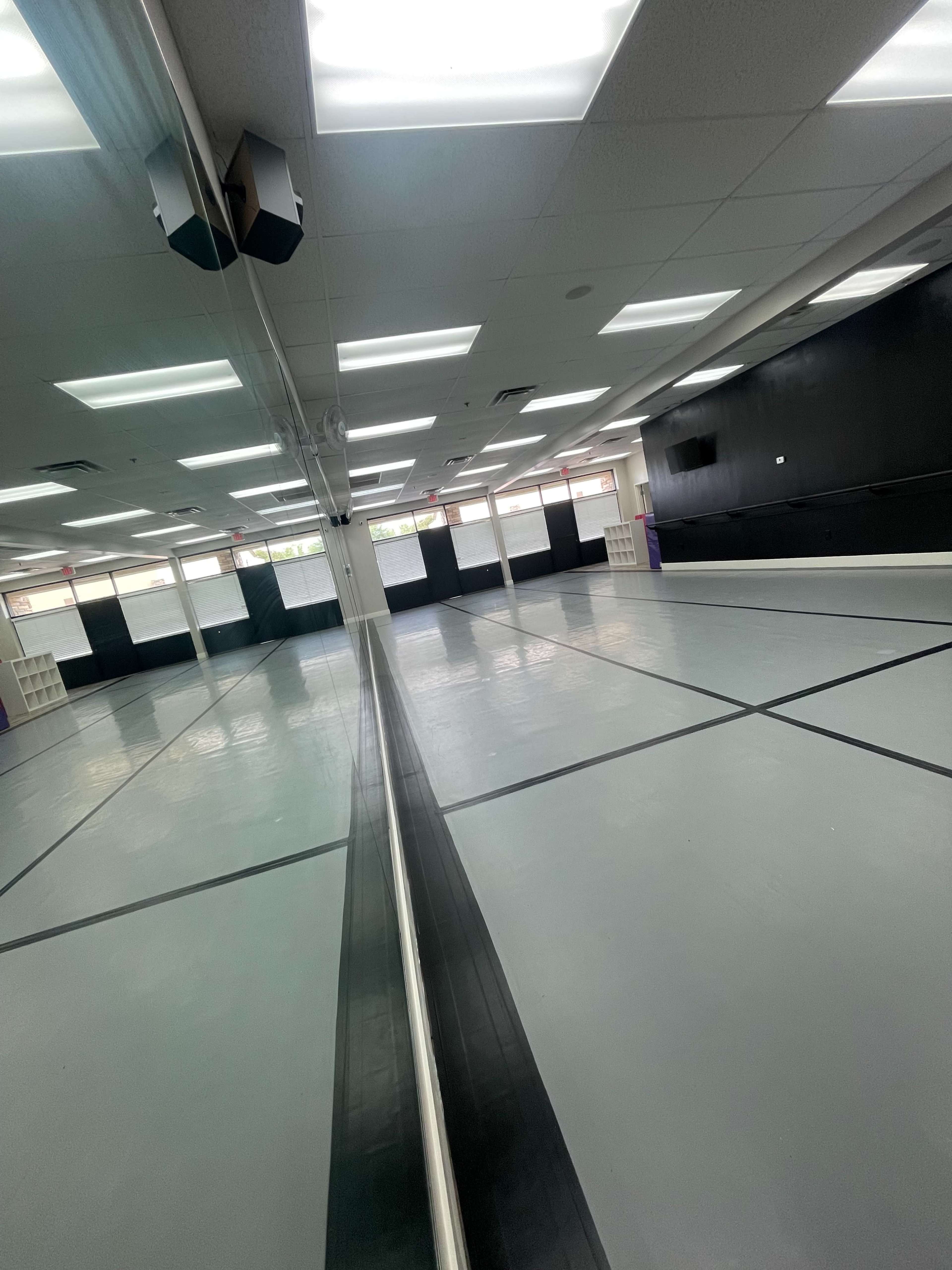 A spacious dance studio with a mirrored wall and gray flooring under bright overhead lights.