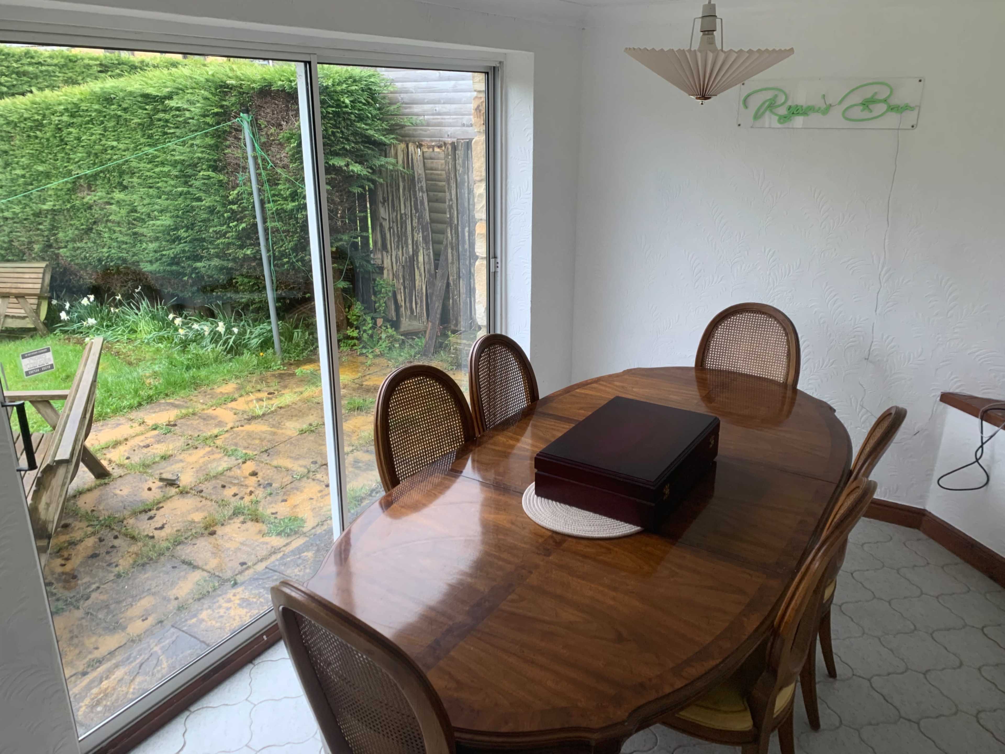 A rectangular wooden dining table with six chairs is situated in a brightly lit room, next to a sliding glass door that opens to a garden with patio stones.