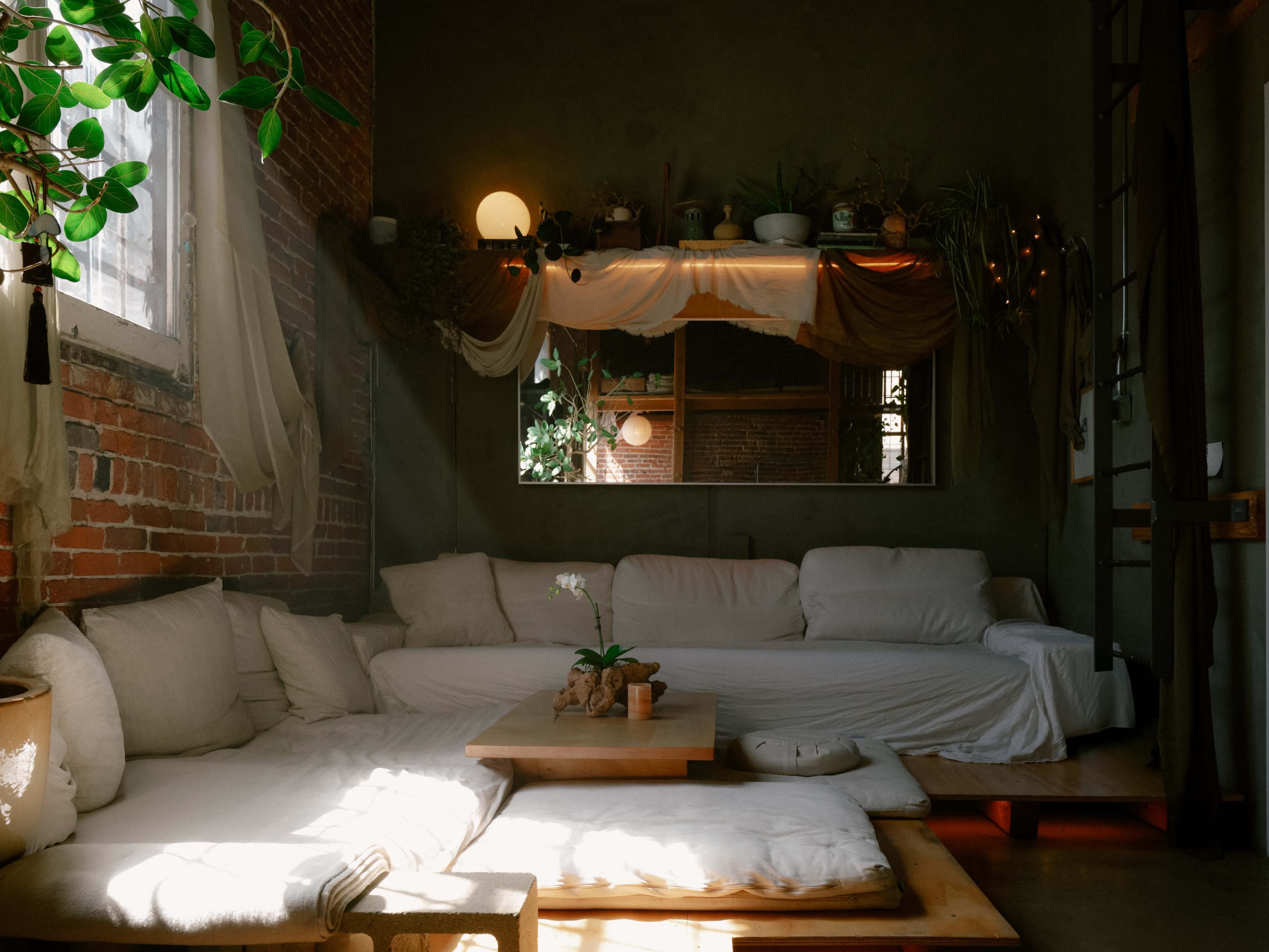 The image features a cozy living space with a large, cushioned seating area, a wooden table, and plants adorning the shelves and windows.