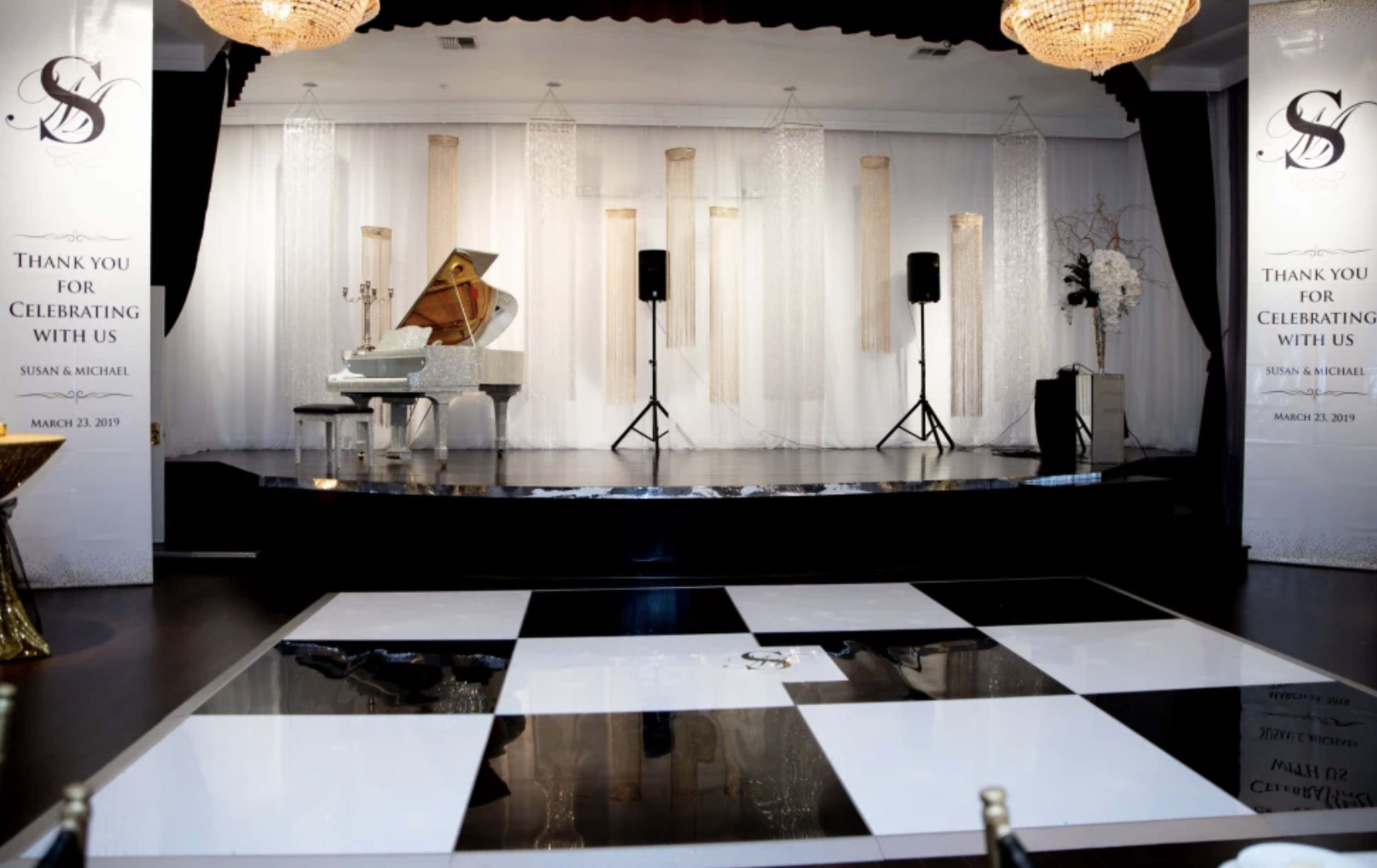 A decorated event stage with a grand piano, speakers, a black and white dance floor, and elegant drapery in the background.