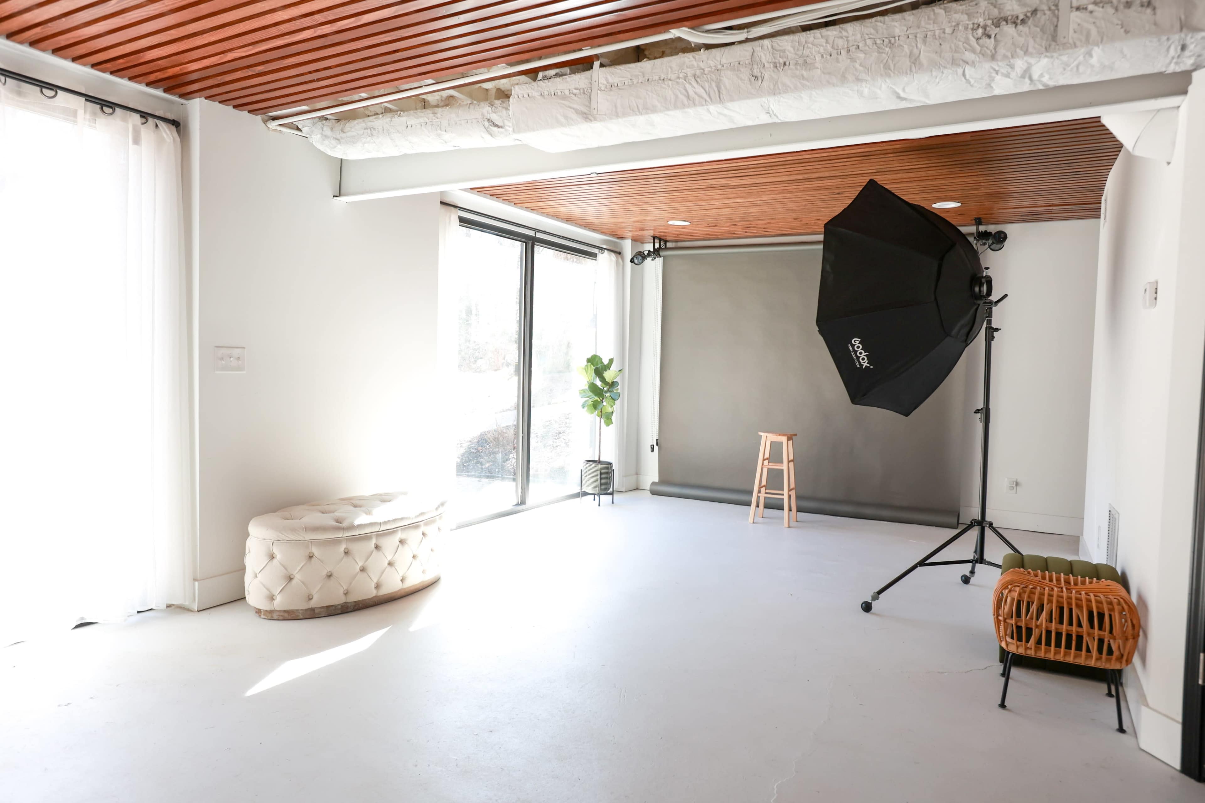 The image shows a bright, uncluttered studio space featuring large windows, a photography backdrop, a stool, and lighting equipment.