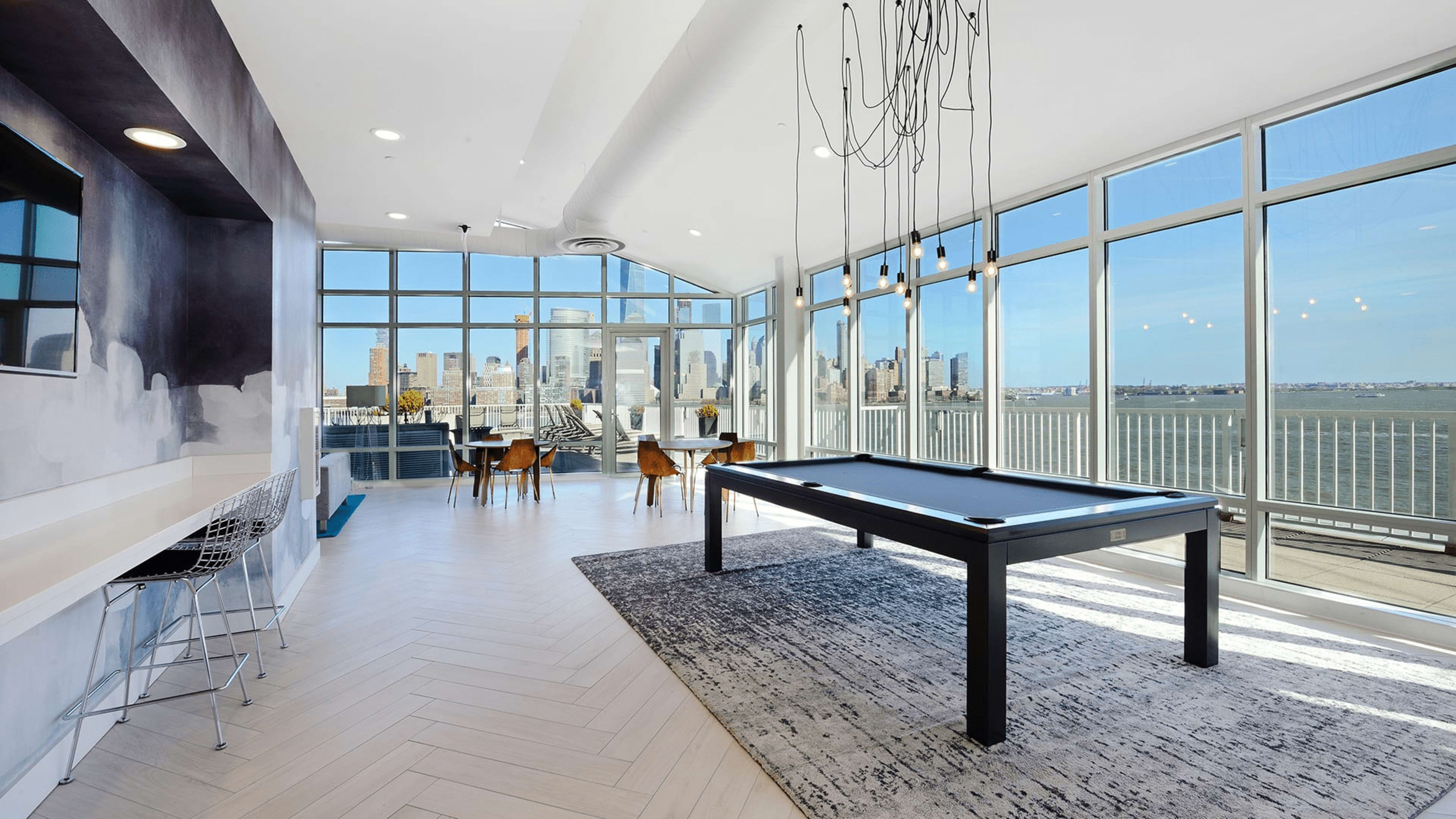 A modern room features a billiards table, stylish seating, and large windows offering a view of the city skyline and water.