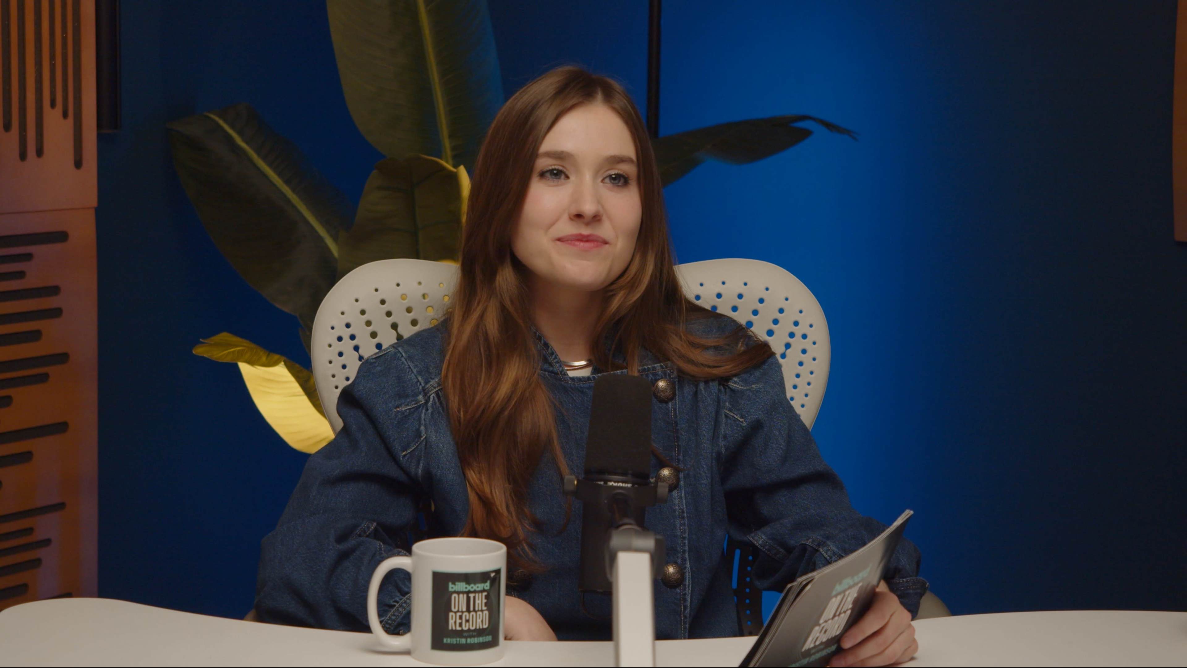 A woman with long brown hair sits in front of a microphone and holds a tablet, with green plants in the background.