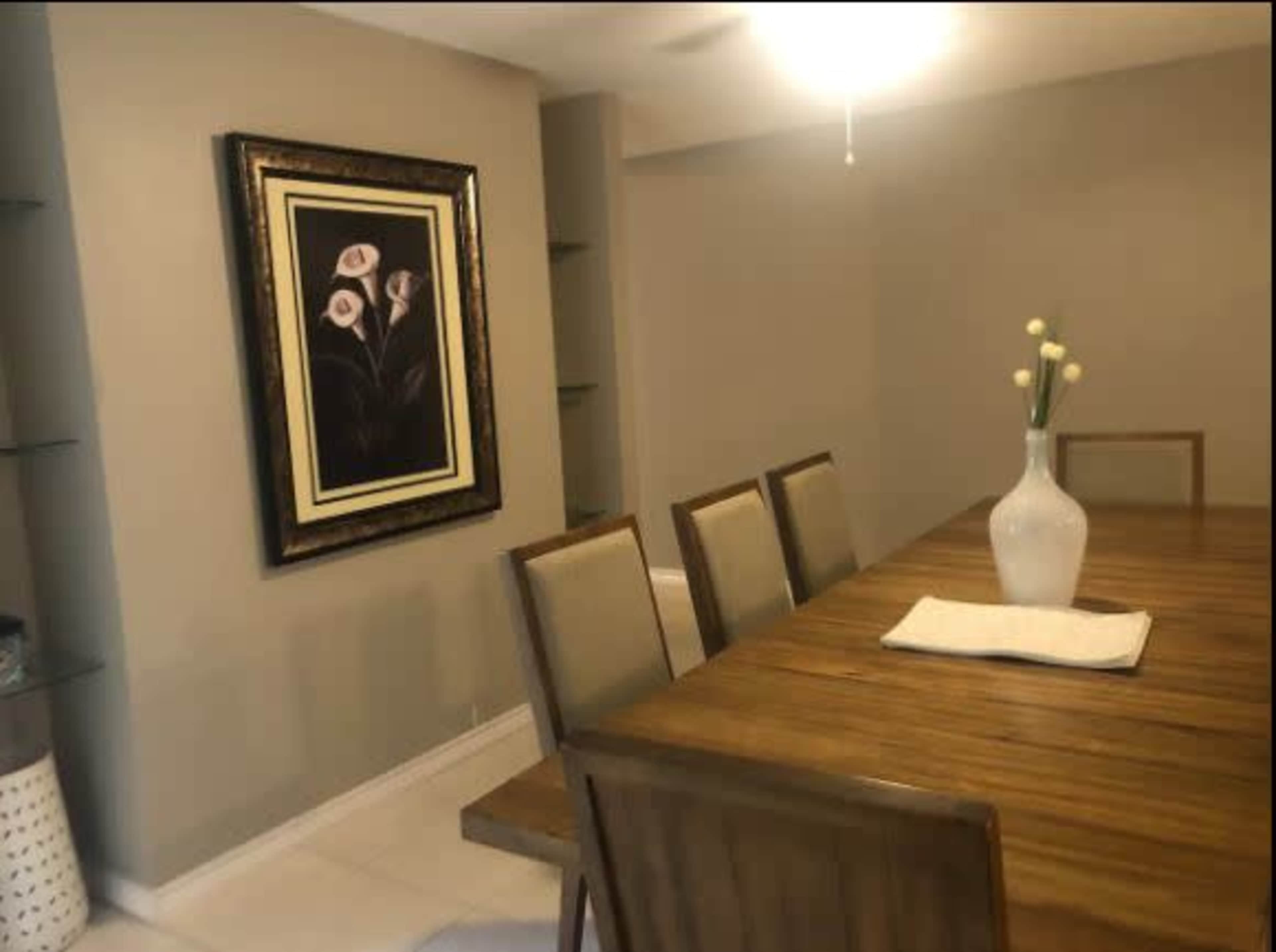 A dining room features a wooden table surrounded by eight chairs, with a large floral painting on the wall and a vase on the table.