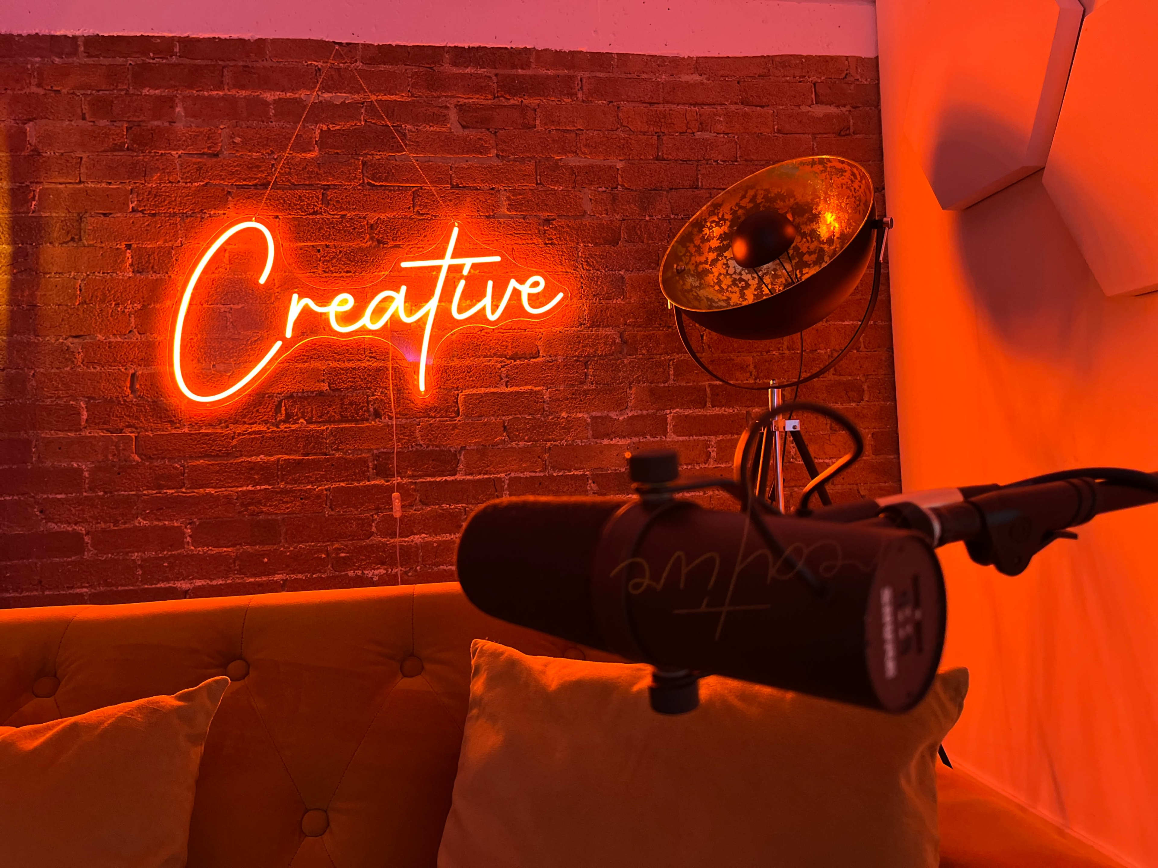 A neon sign reading "Creative" is illuminated against a brick wall, with a microphone positioned in the foreground and a vintage lamp in the background.