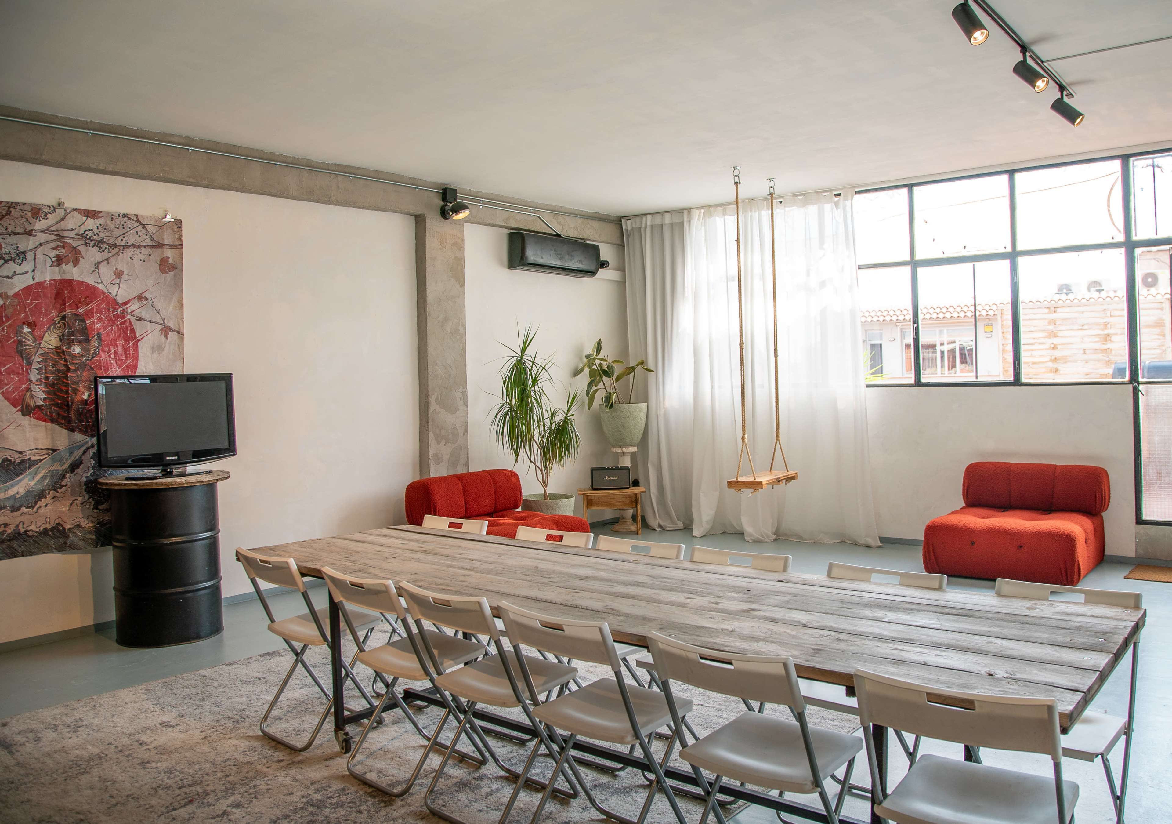 The image shows a modern, minimalist meeting room with a large wooden table surrounded by white folding chairs, a television, and two red sofas.