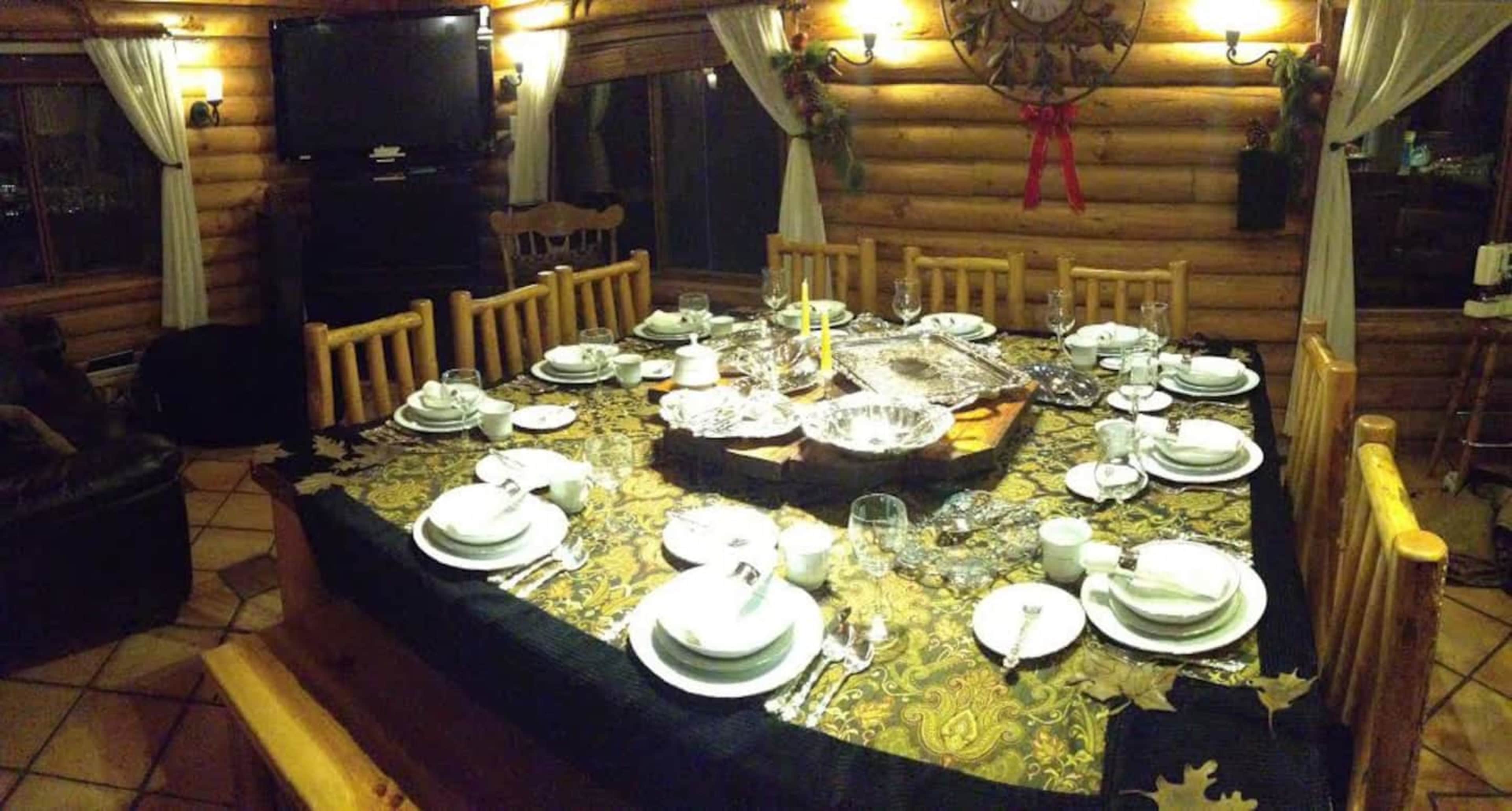 A large, neatly set dining table is arranged in a log cabin, featuring white dishware and a decorative tablecloth, with a view of wooden walls and a television in the background.