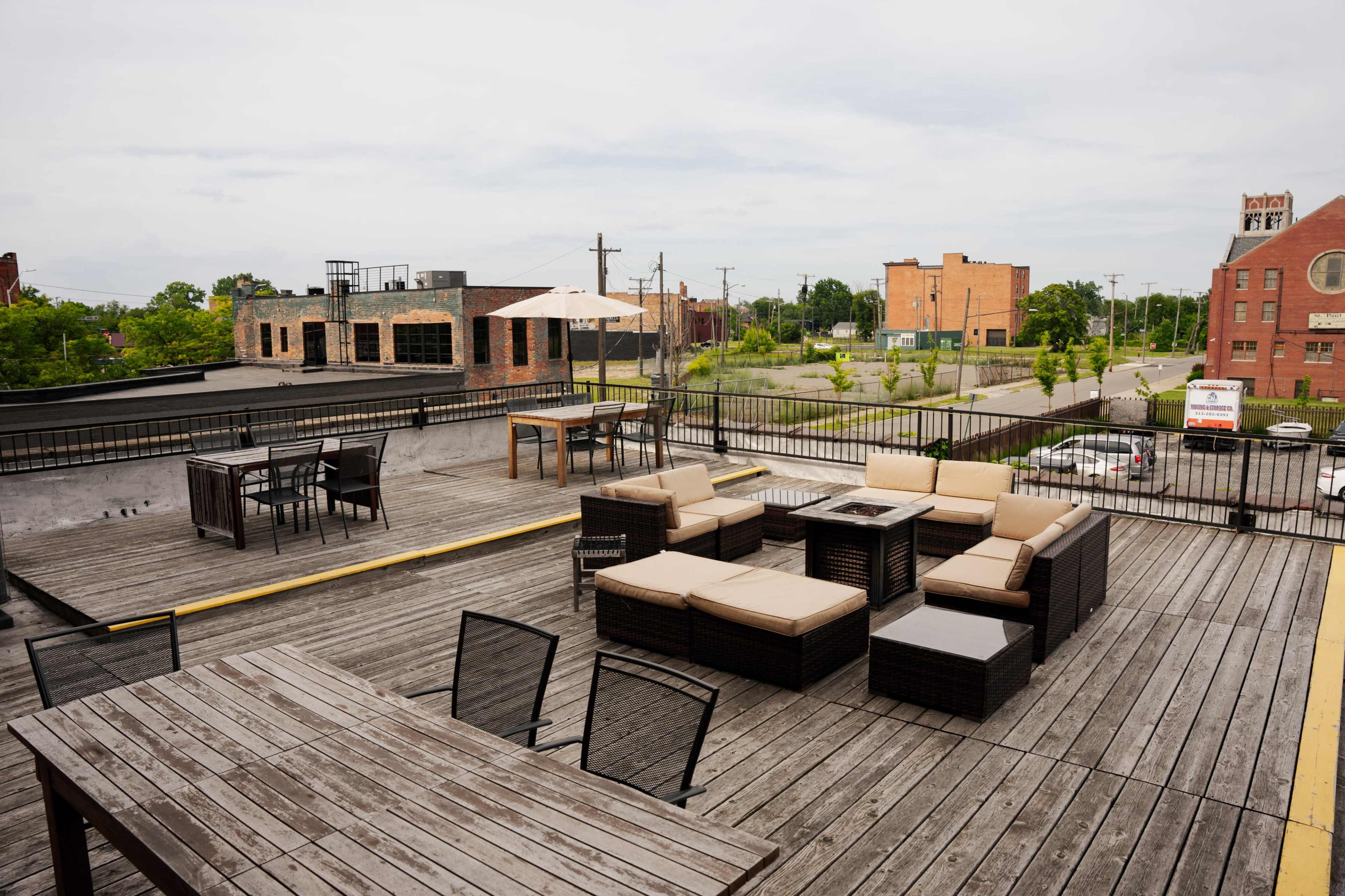 A rooftop terrace with wooden decking, featuring several seating arrangements and tables surrounded by buildings in an urban area.