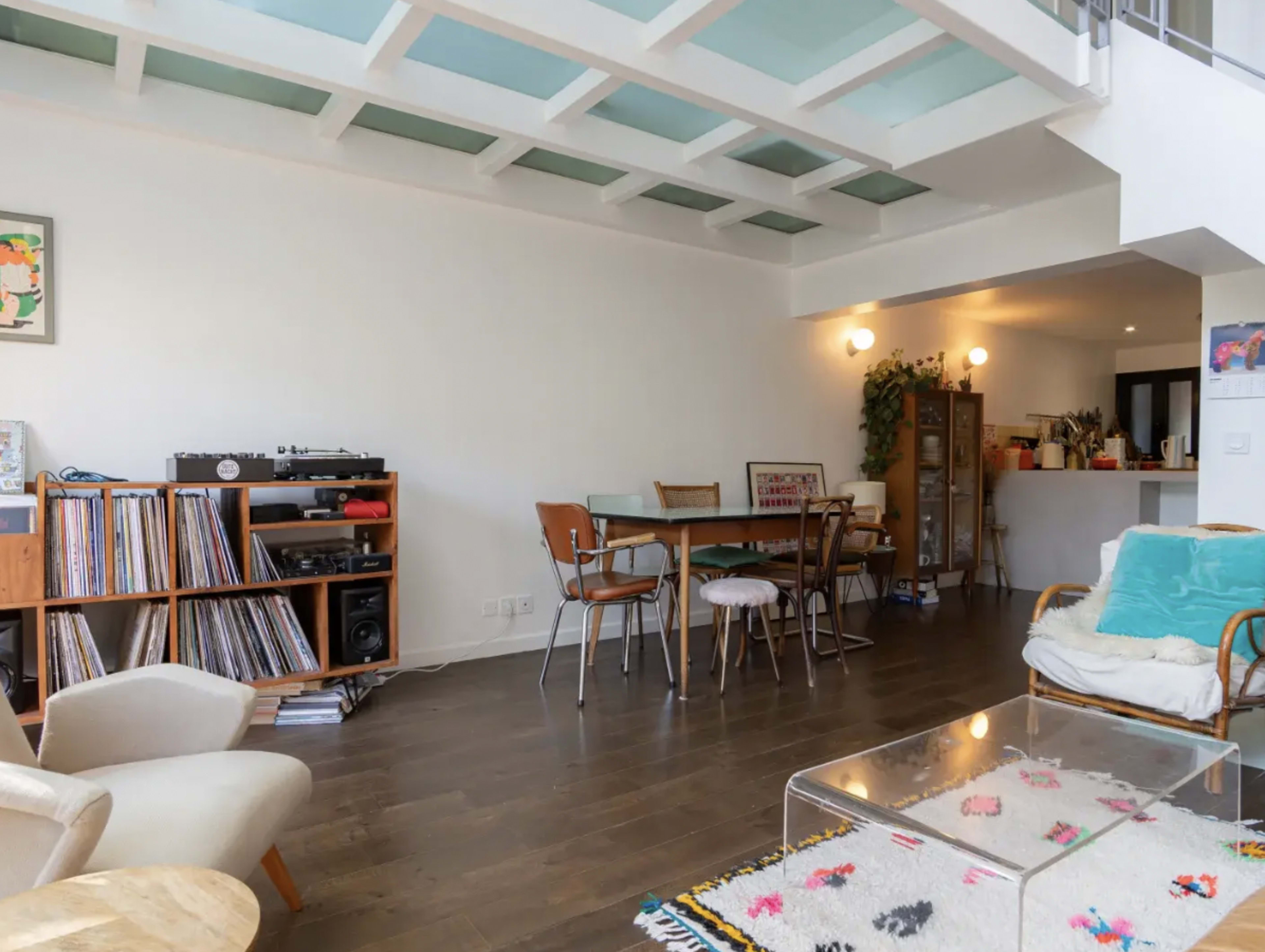 The image features a modern living space with wooden flooring, a dining table, a record player, and a mix of chairs and decor.
