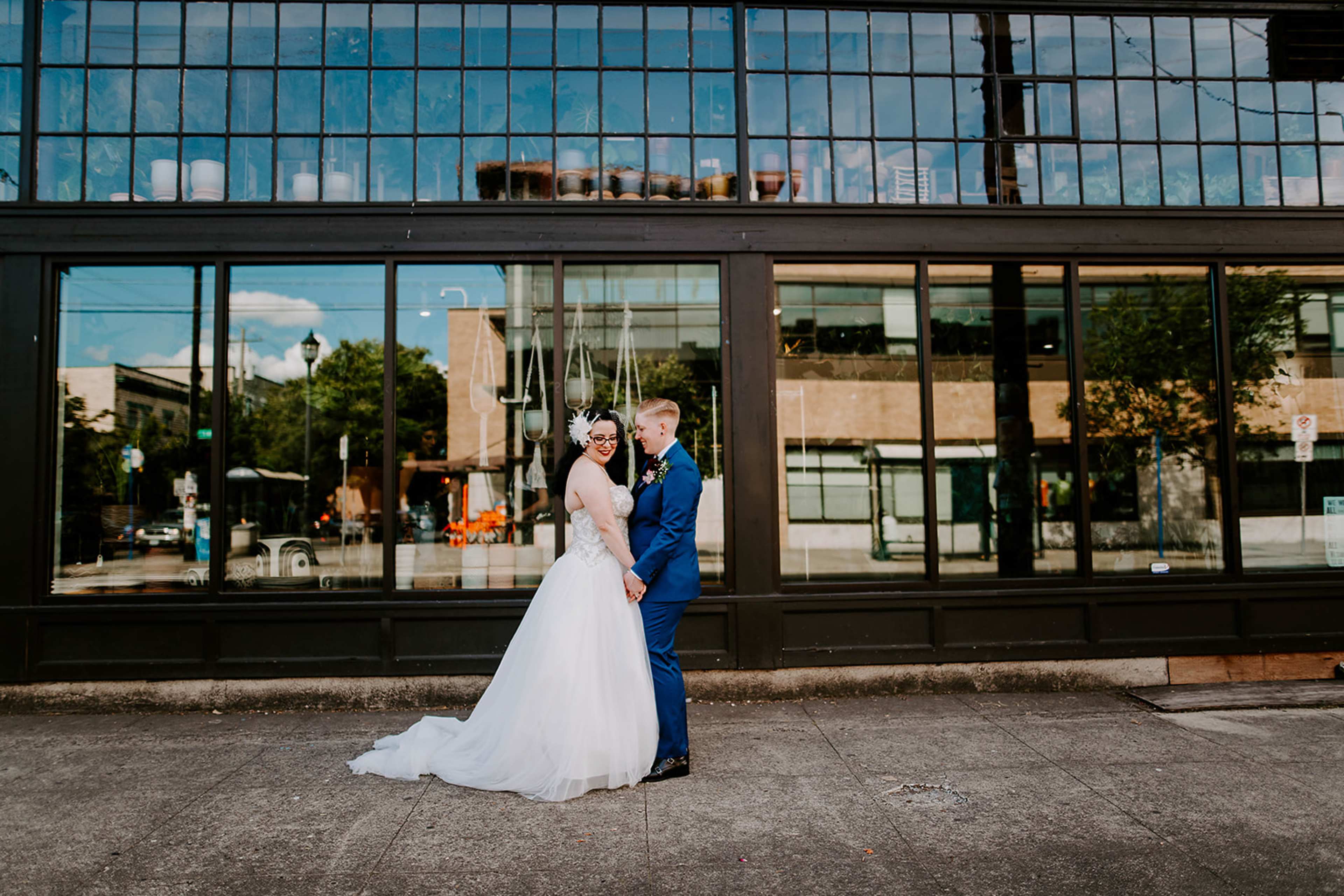 A bride in a white gown and a groom in a blue suit hold hands while standing in front of a large glass window reflecting the urban surroundings.