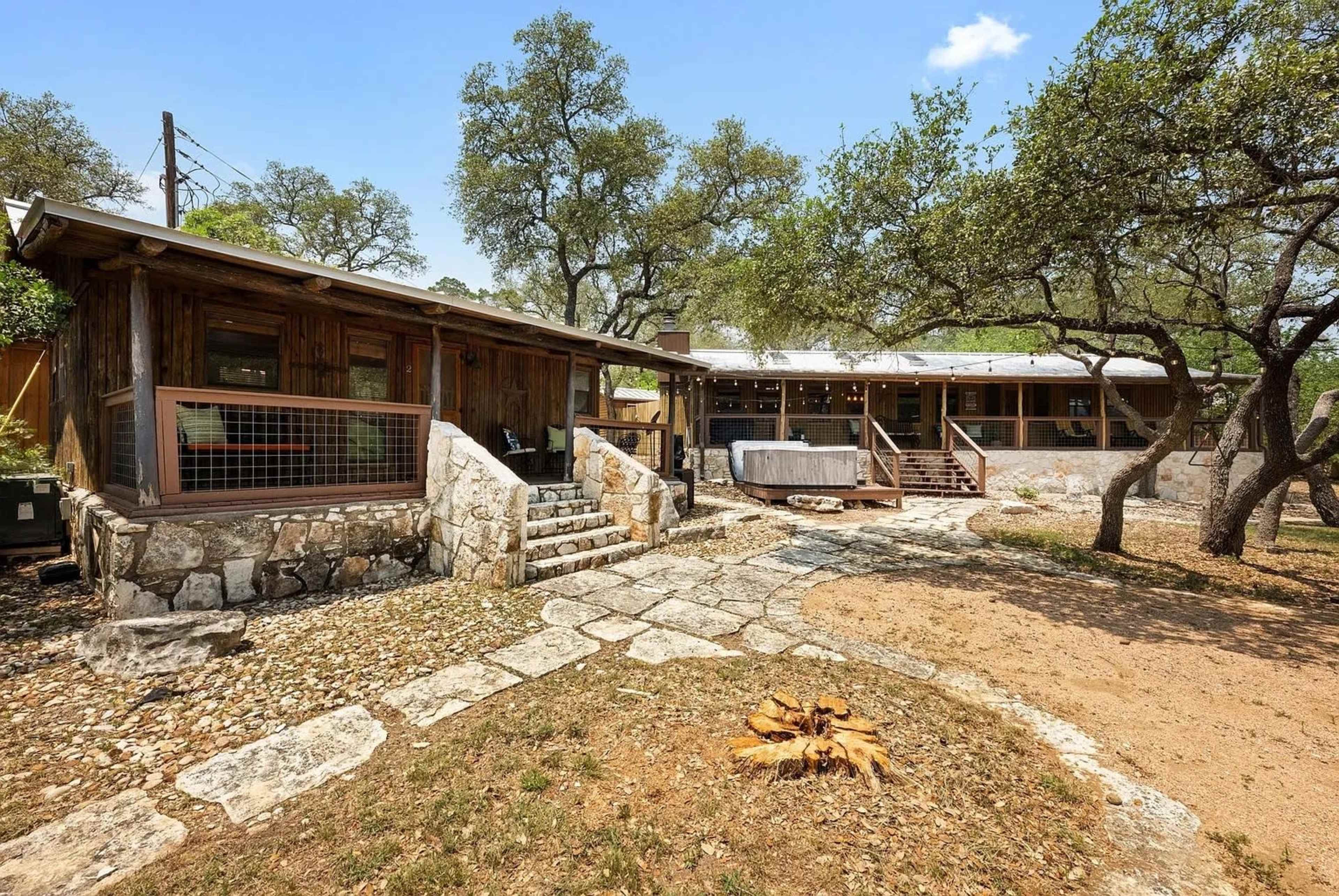 The image shows a rustic cabin complex with stone pathways, surrounded by trees and a sandy area.
