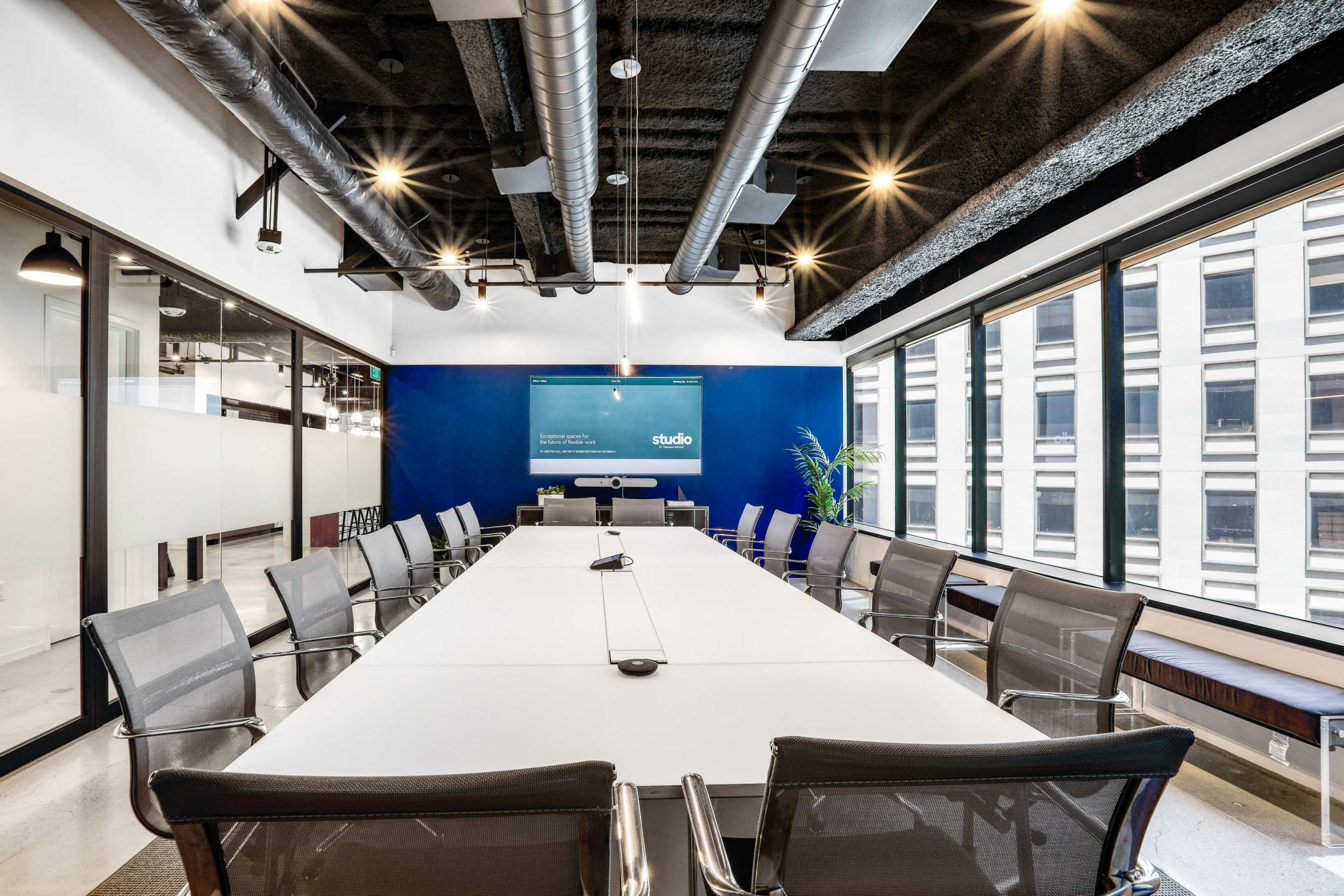 A modern conference room features a long white table surrounded by metal chairs, with large windows offering views of a cityscape.