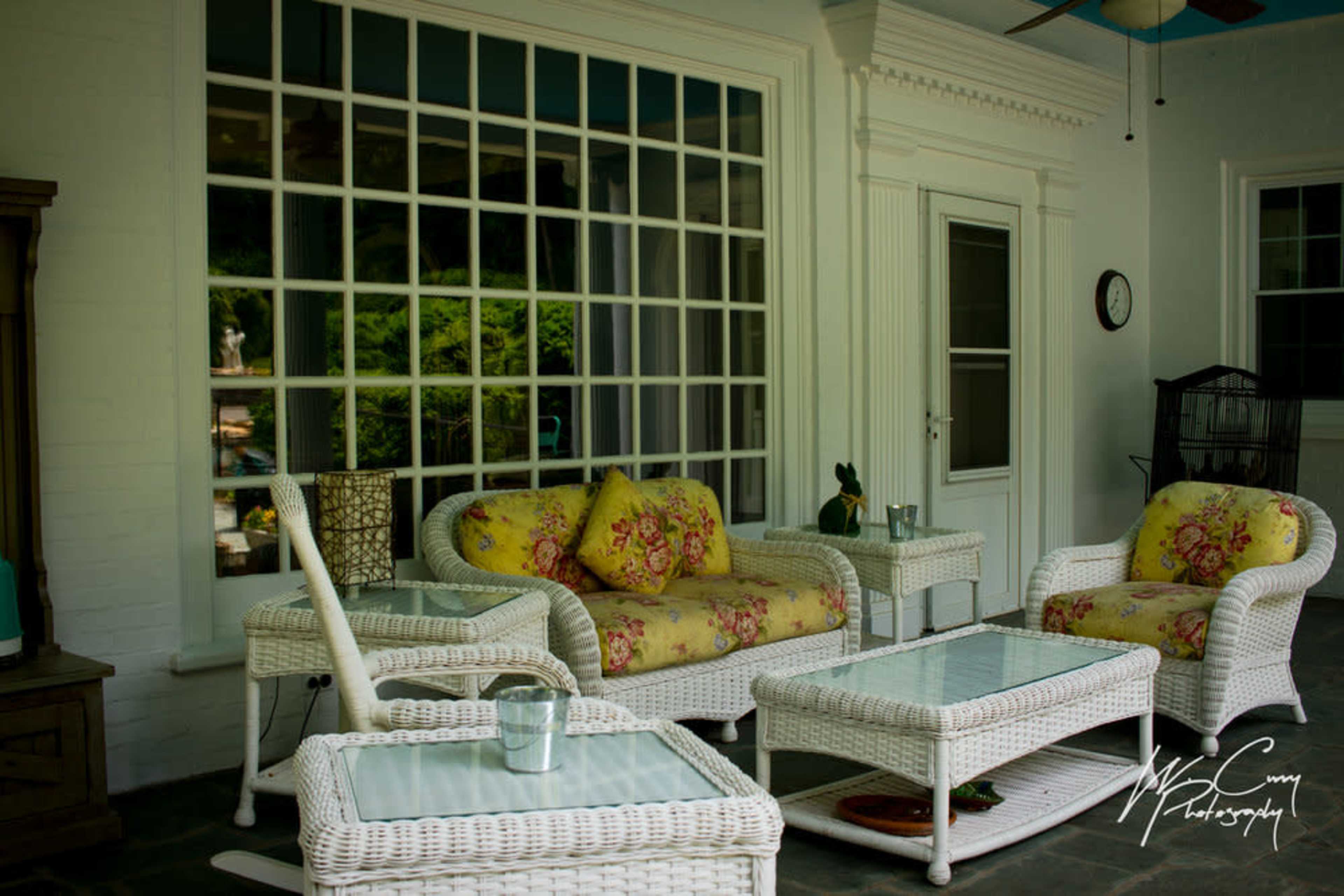 A porch area with white wicker furniture, including two chairs and a coffee table, arranged around a glass-topped table, with a large window in the background.