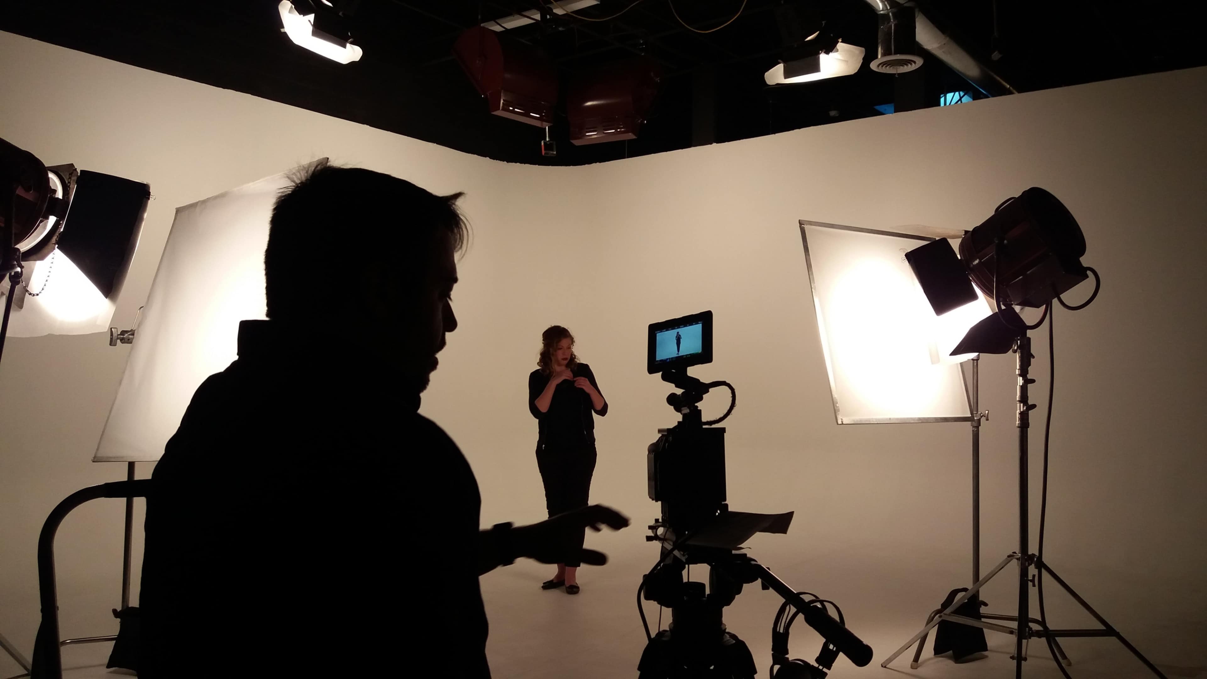 A person operates a camera while another stands in front of lighting equipment in a photography studio.