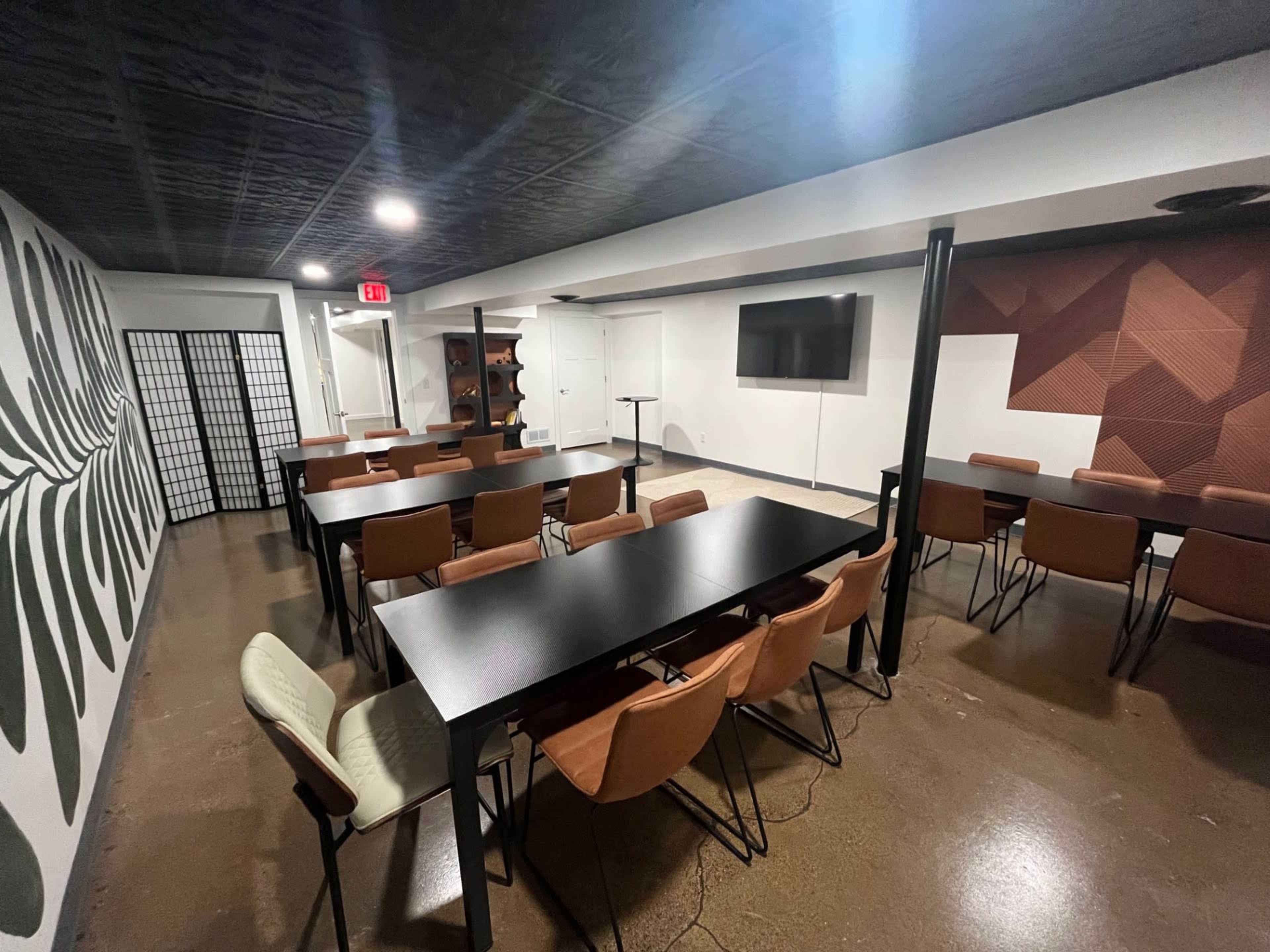 The image shows a modern meeting room with multiple tables and chairs, a wall-mounted TV, and decorative wall panels.