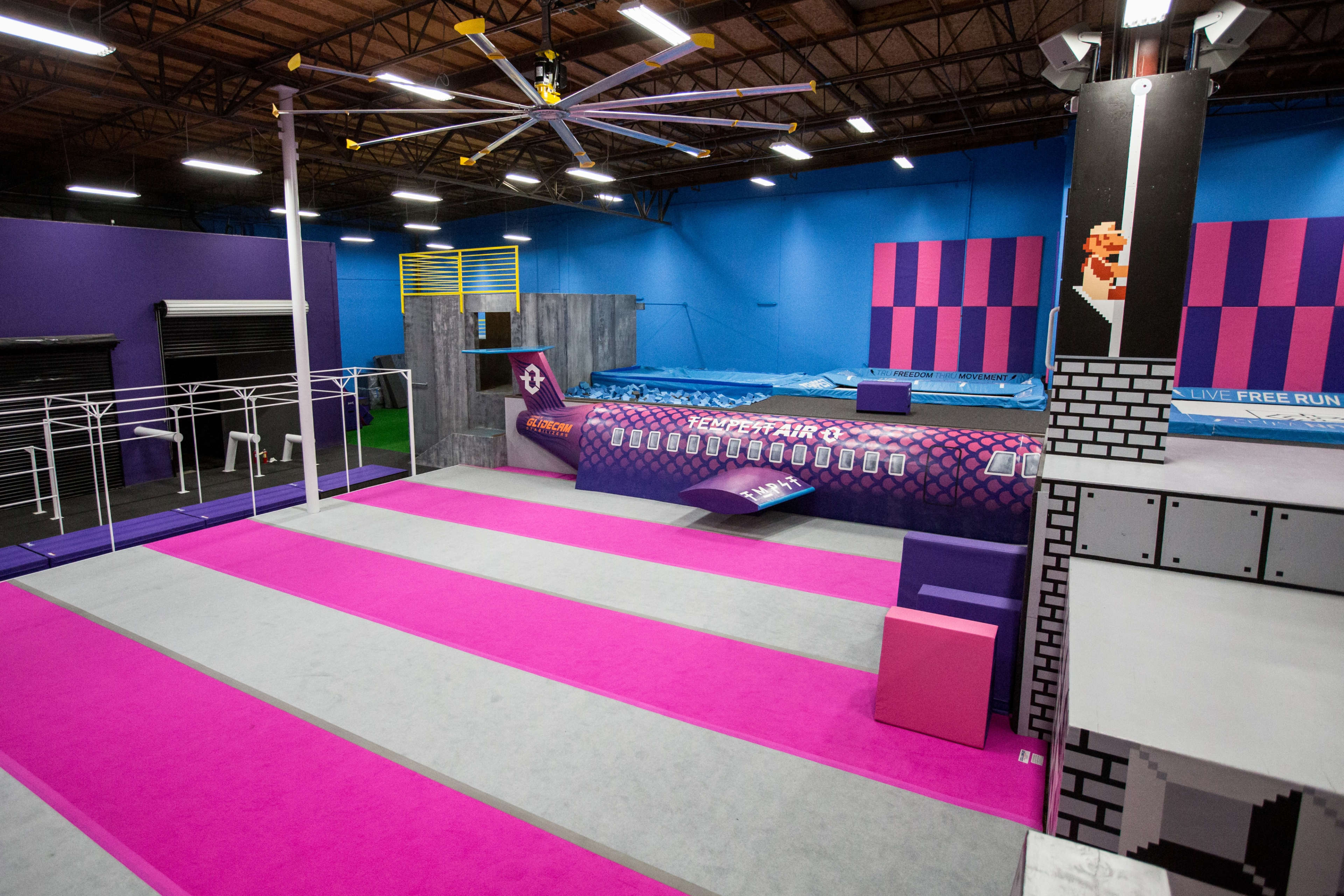 An indoor parkour gym with colorful walls, various obstacles, and a large foam pit at one end.