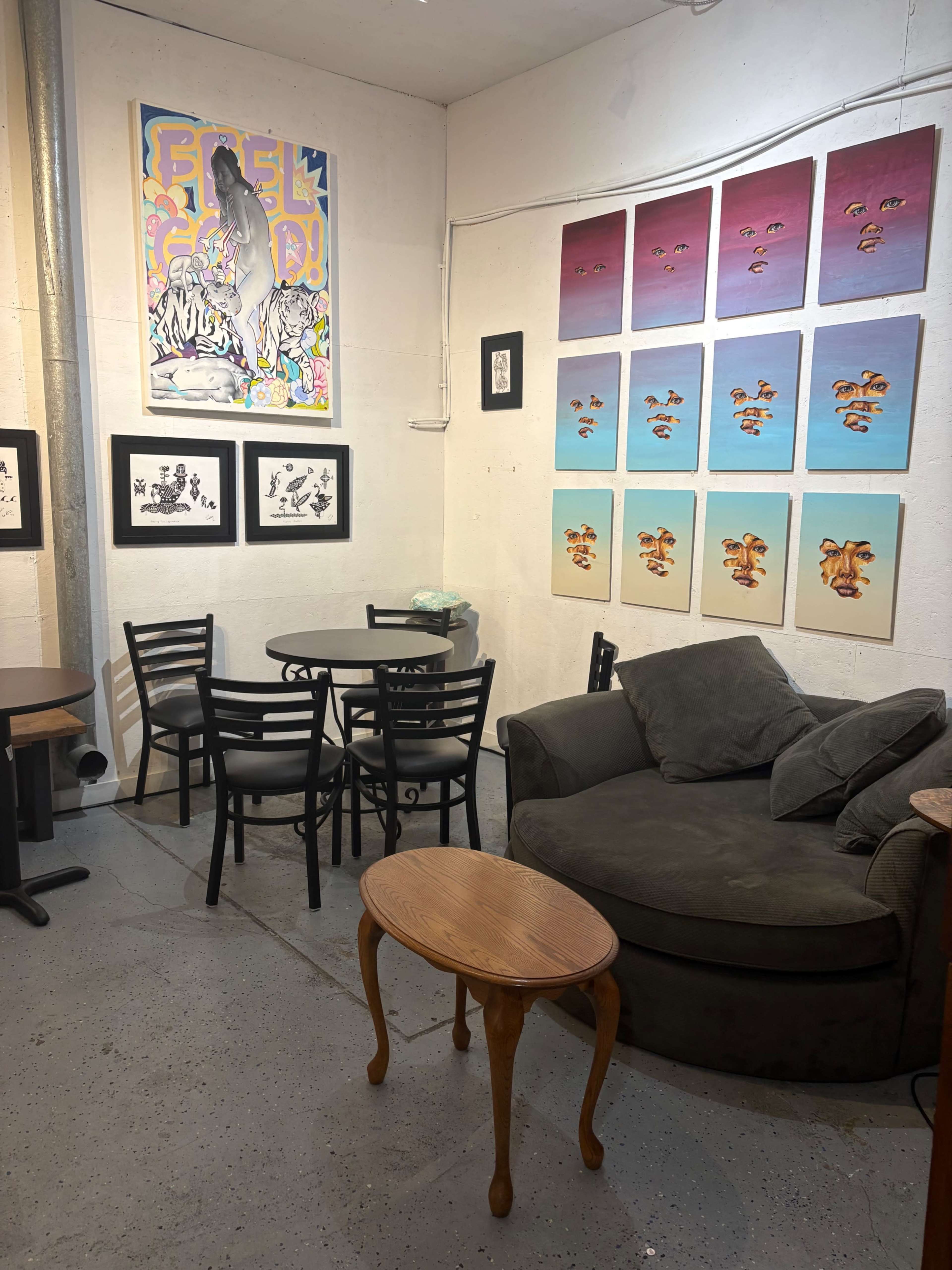The image shows a cozy corner of an art space featuring a round table with four chairs, a dark green couch, and several framed artworks on the walls.