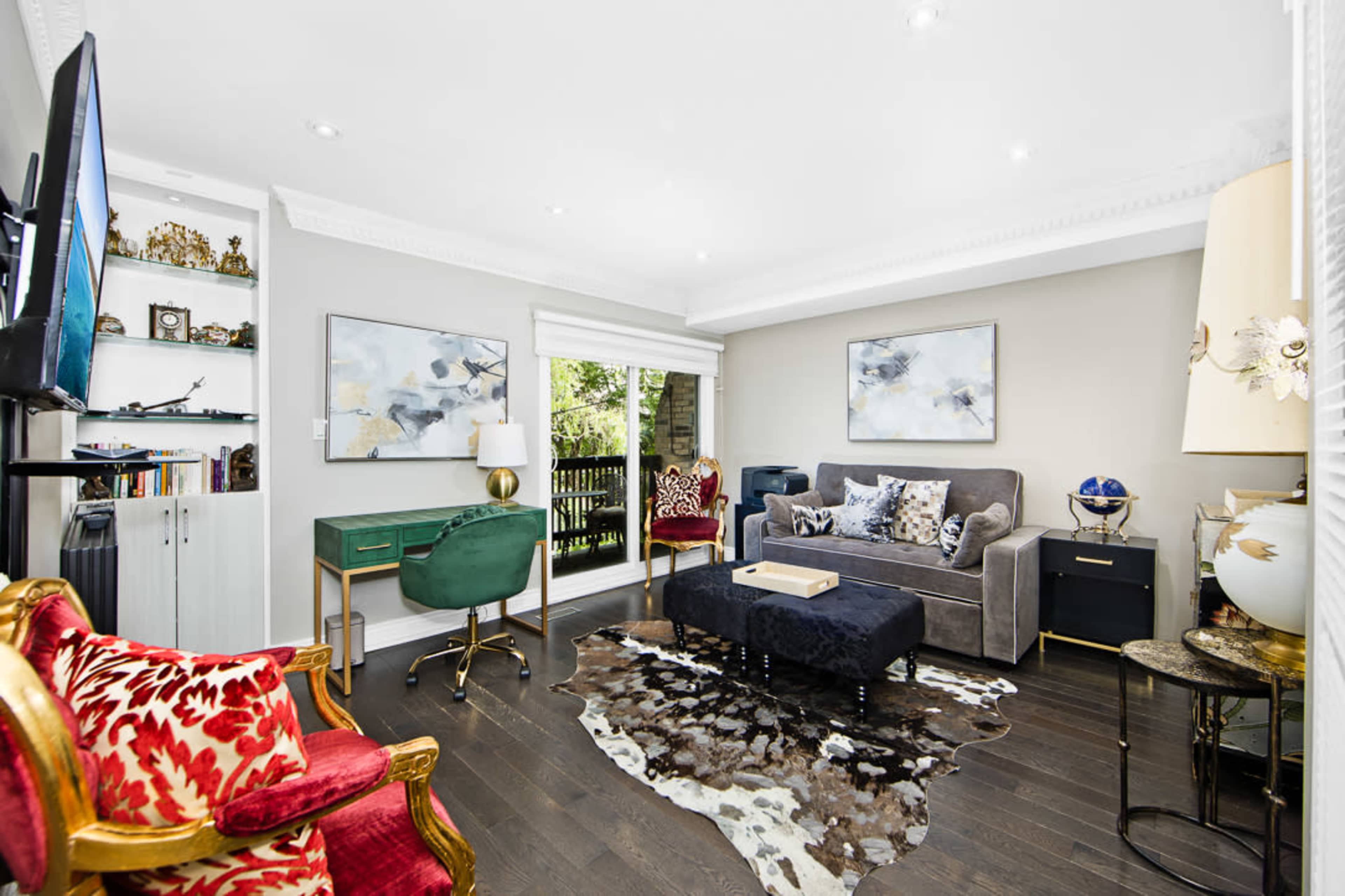 A stylish living room features a gray sofa, green desk, patterned rug, and large windows with natural light.