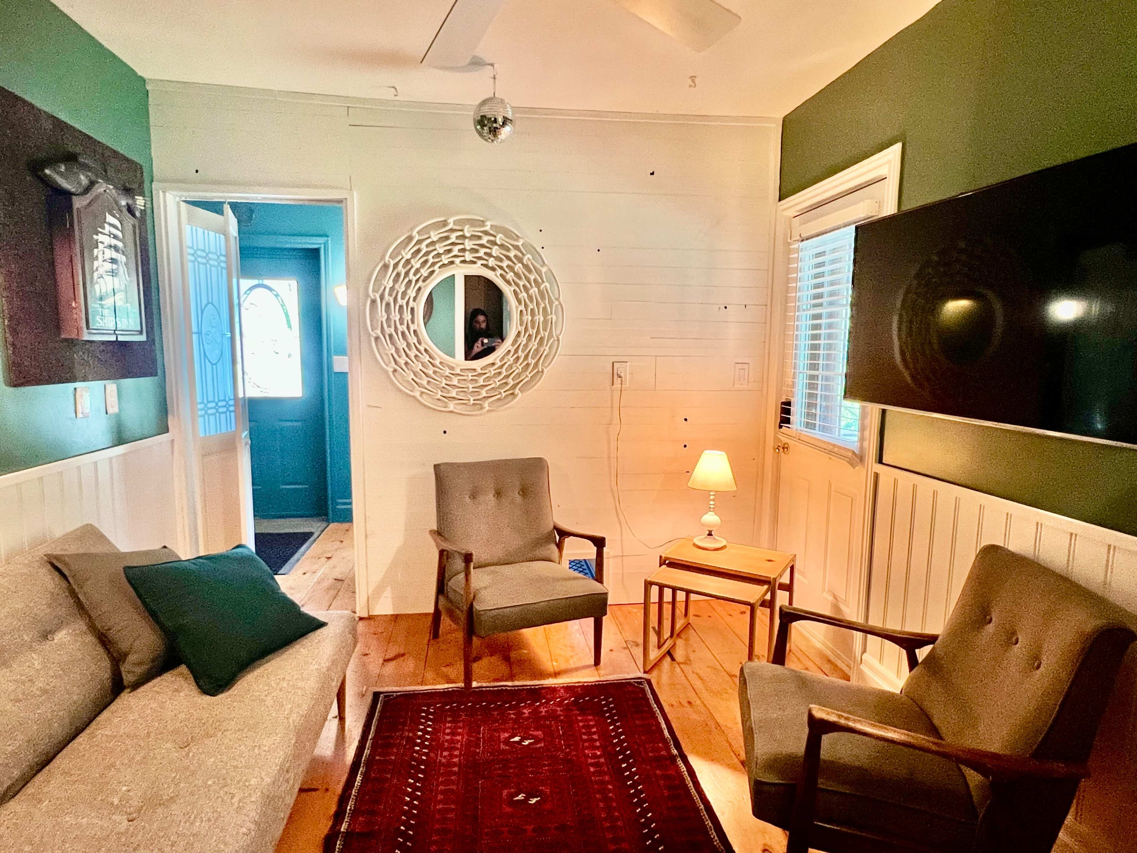 A cozy living room with a gray sofa and two armchairs facing a wooden side table and a wall-mounted television, while a mirror and a door lead to another room.