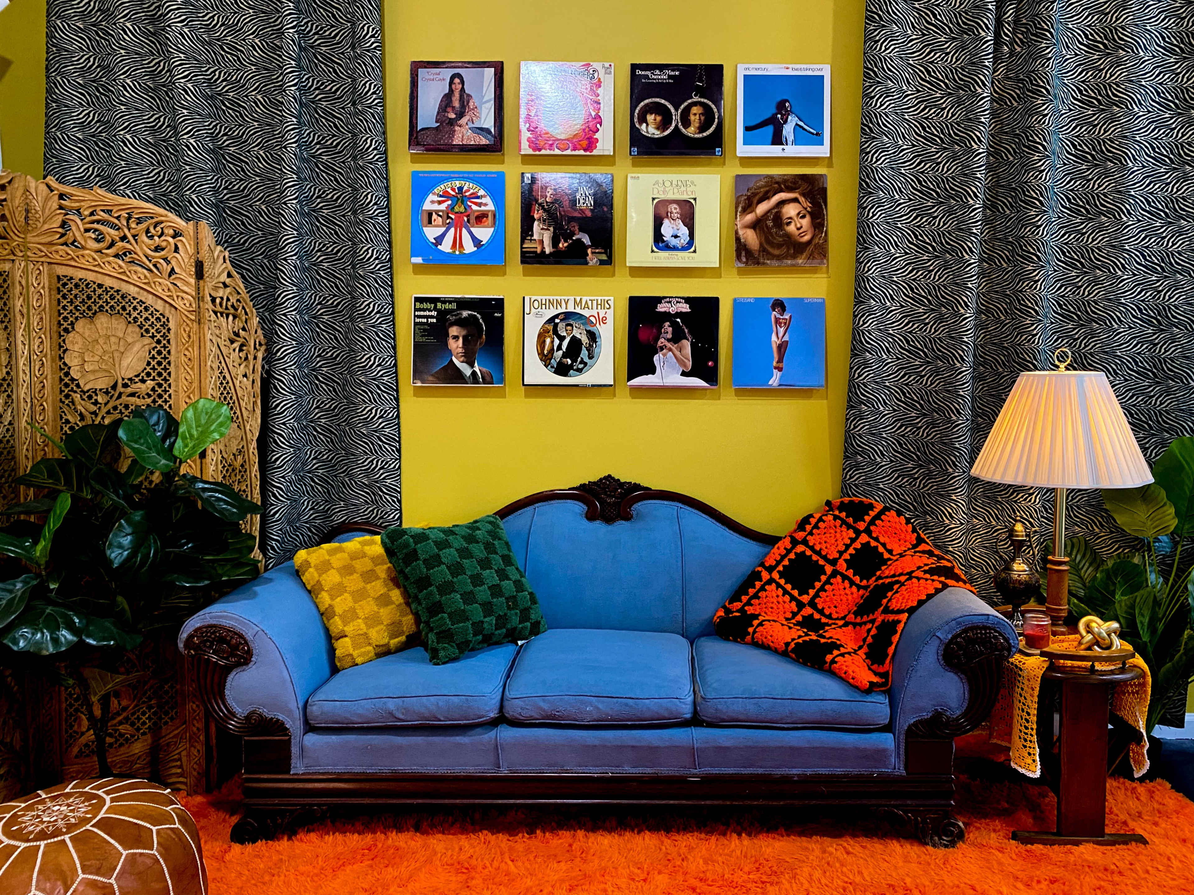 A blue couch with patterned pillows is positioned against a bright yellow wall adorned with framed vinyl record covers, while indoor plants and a lamp complete the cozy living space.