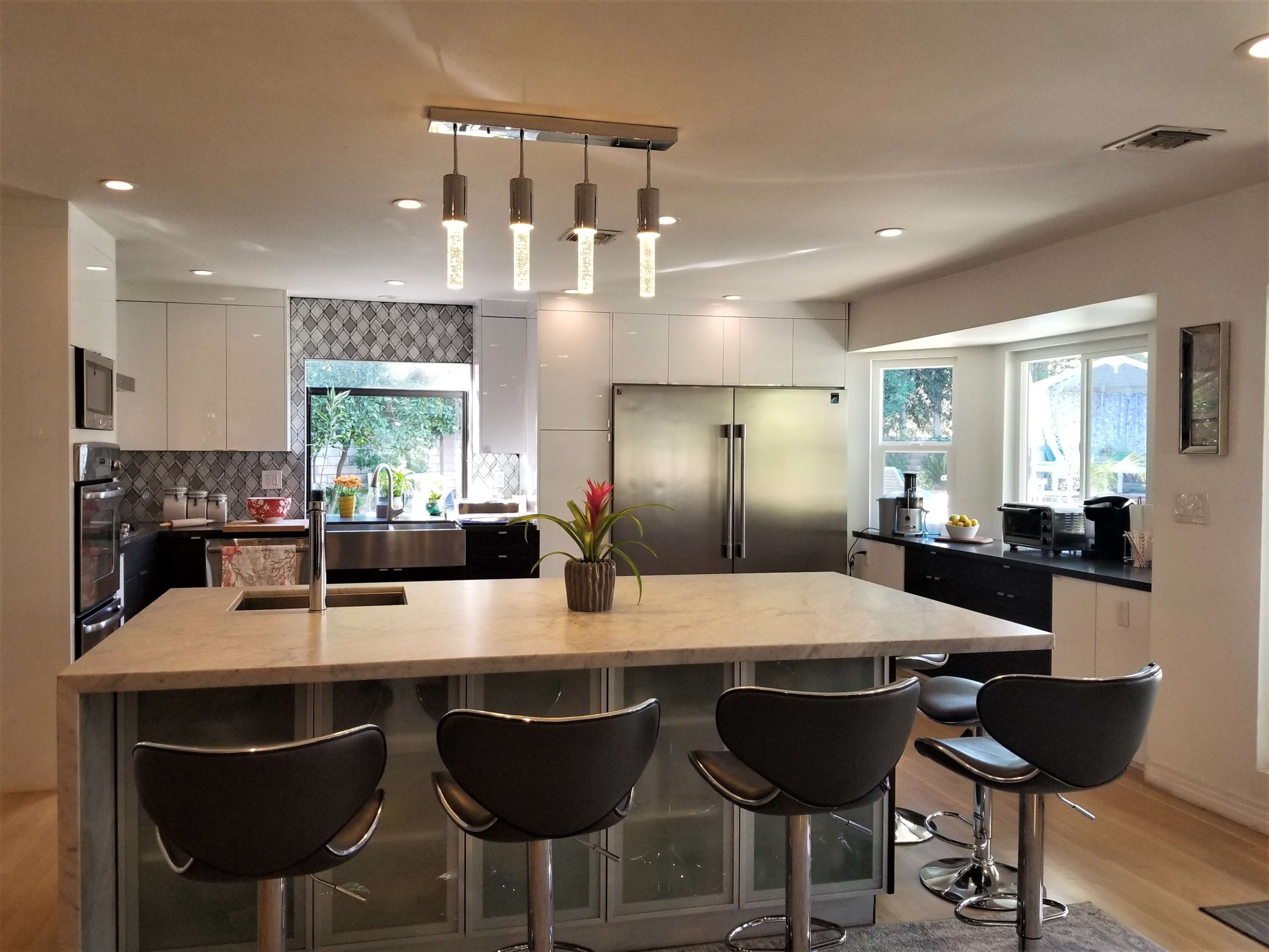 A modern kitchen features a large island with a marble countertop, four black stools, and stainless steel appliances, including a refrigerator and a double oven.