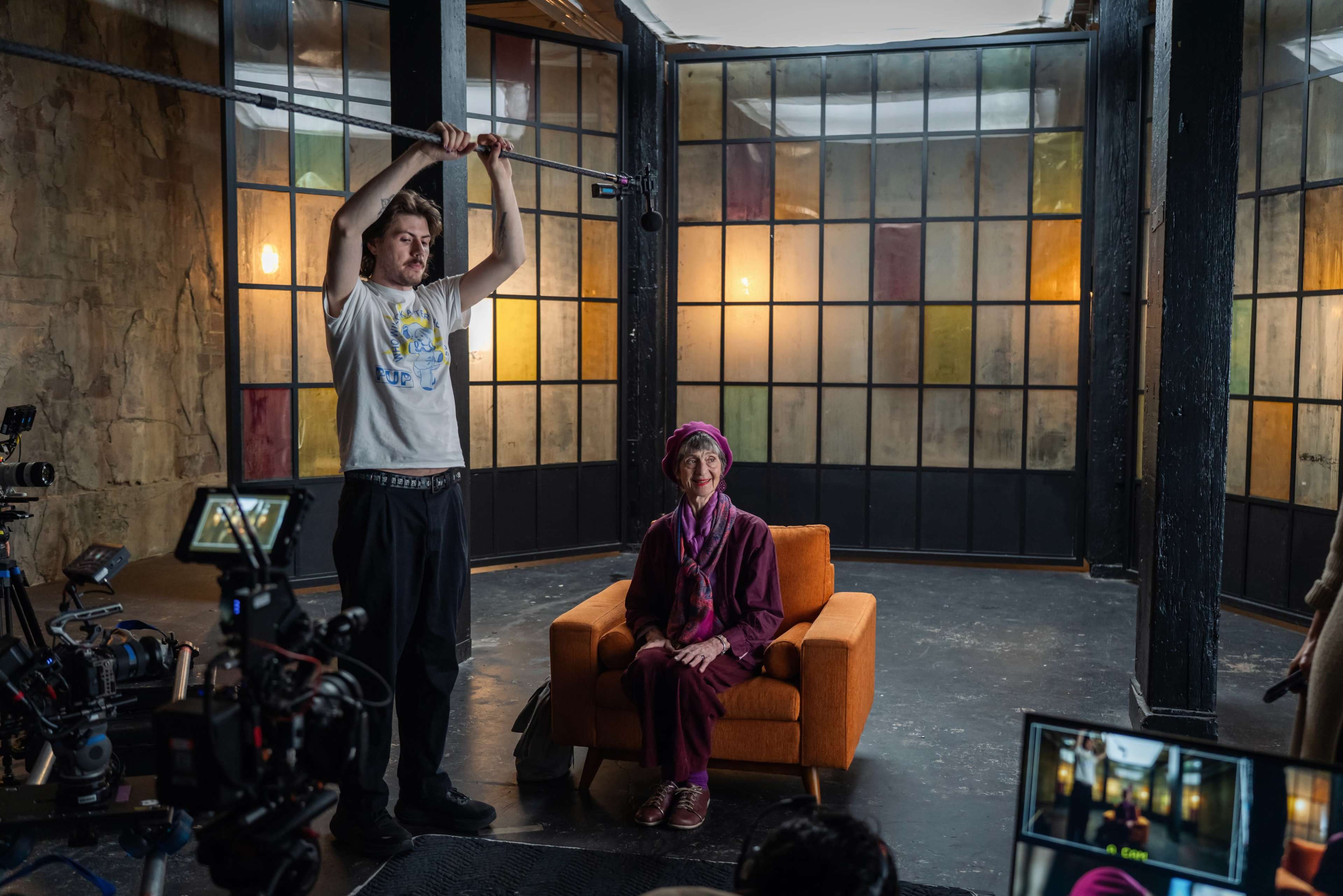 A film crew prepares for a shoot, featuring a woman in a purple outfit sitting on an orange couch while a man holds a microphone above her.