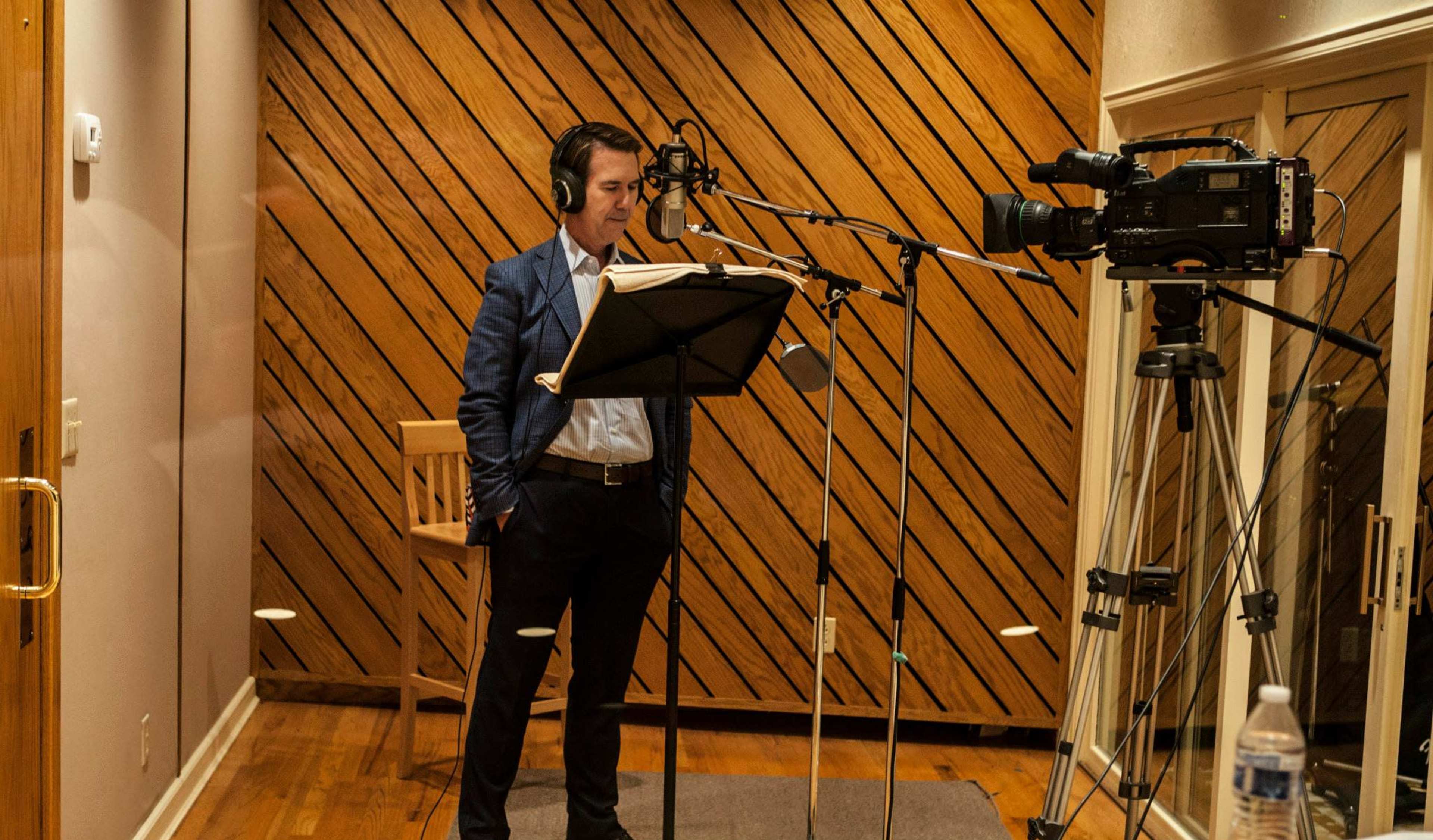 A man in a blue suit stands in a recording studio, wearing headphones and reading from a script while a camera is positioned nearby.