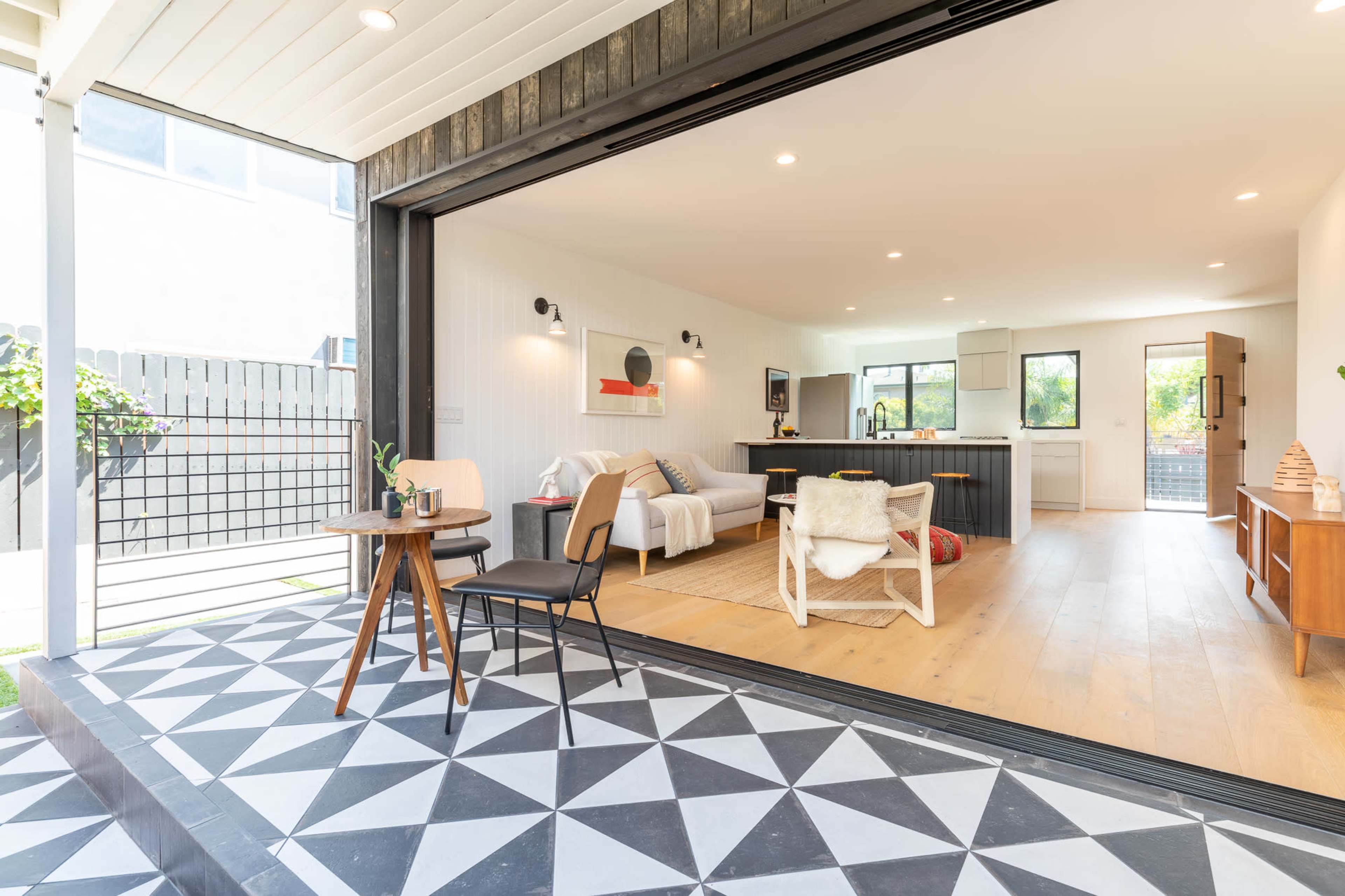The image shows a modern living space with an open floor plan, featuring a seating area, kitchen, and a patio that opens up to a backyard.