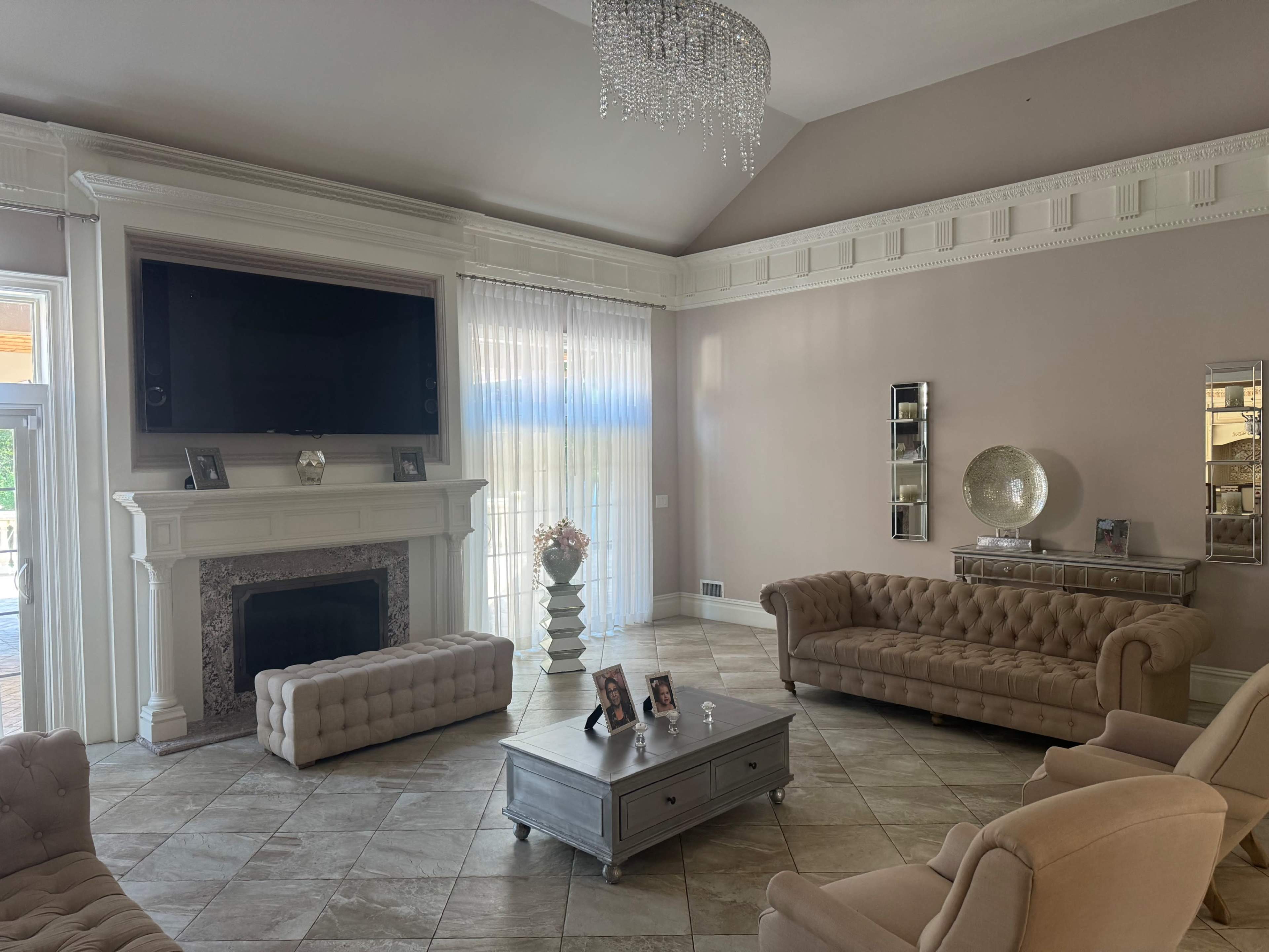 A spacious living room features a large TV above a decorative fireplace, flanked by beige sofas and a glass coffee table set on tiled flooring.