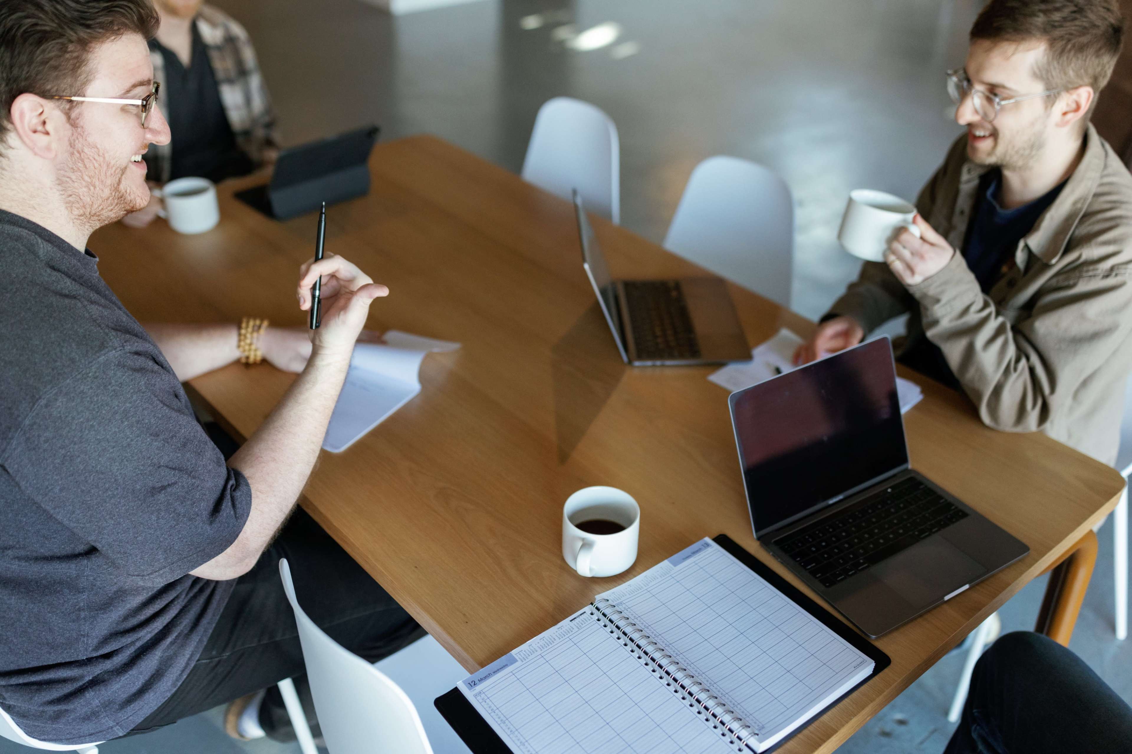 A group of four people is seated around a wooden table, engaged in a discussion while using laptops and holding coffee mugs.