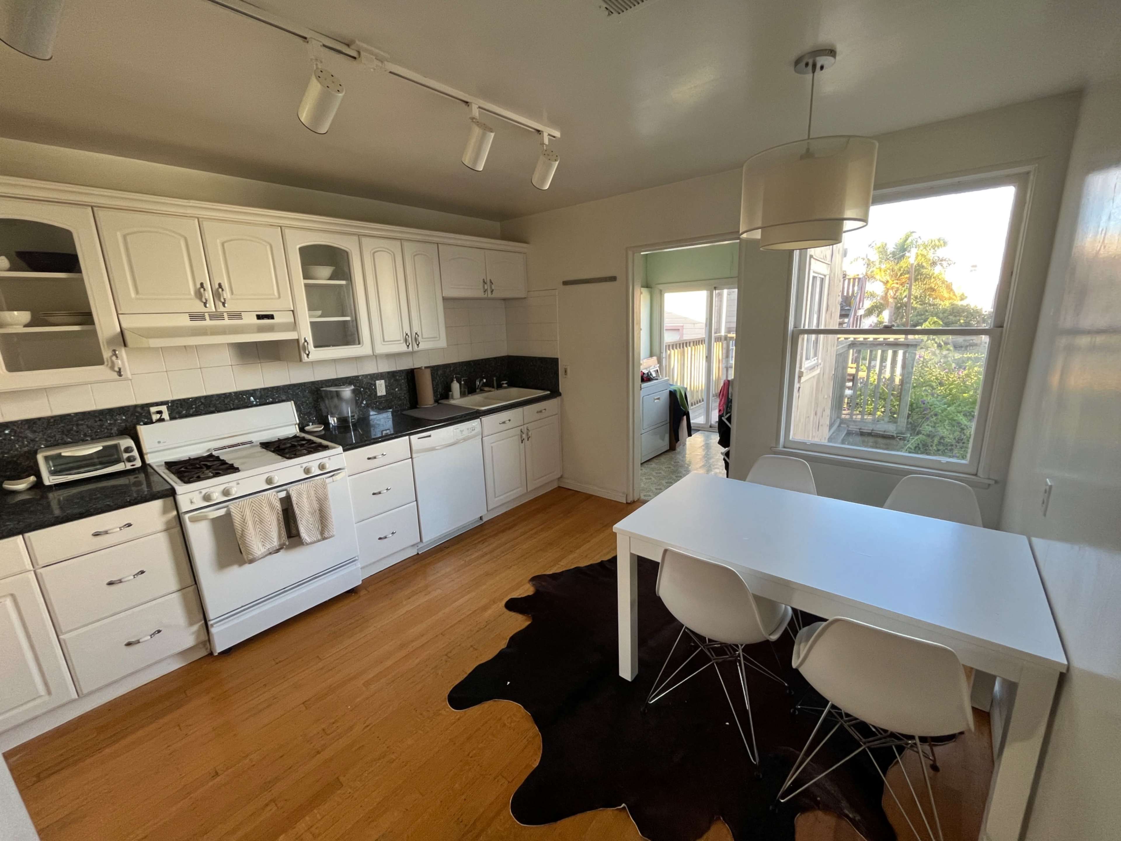 The image shows a kitchen with white cabinets, a gas stove, a dishwasher, and a dining table with modern chairs, illuminated by natural light from a nearby window.