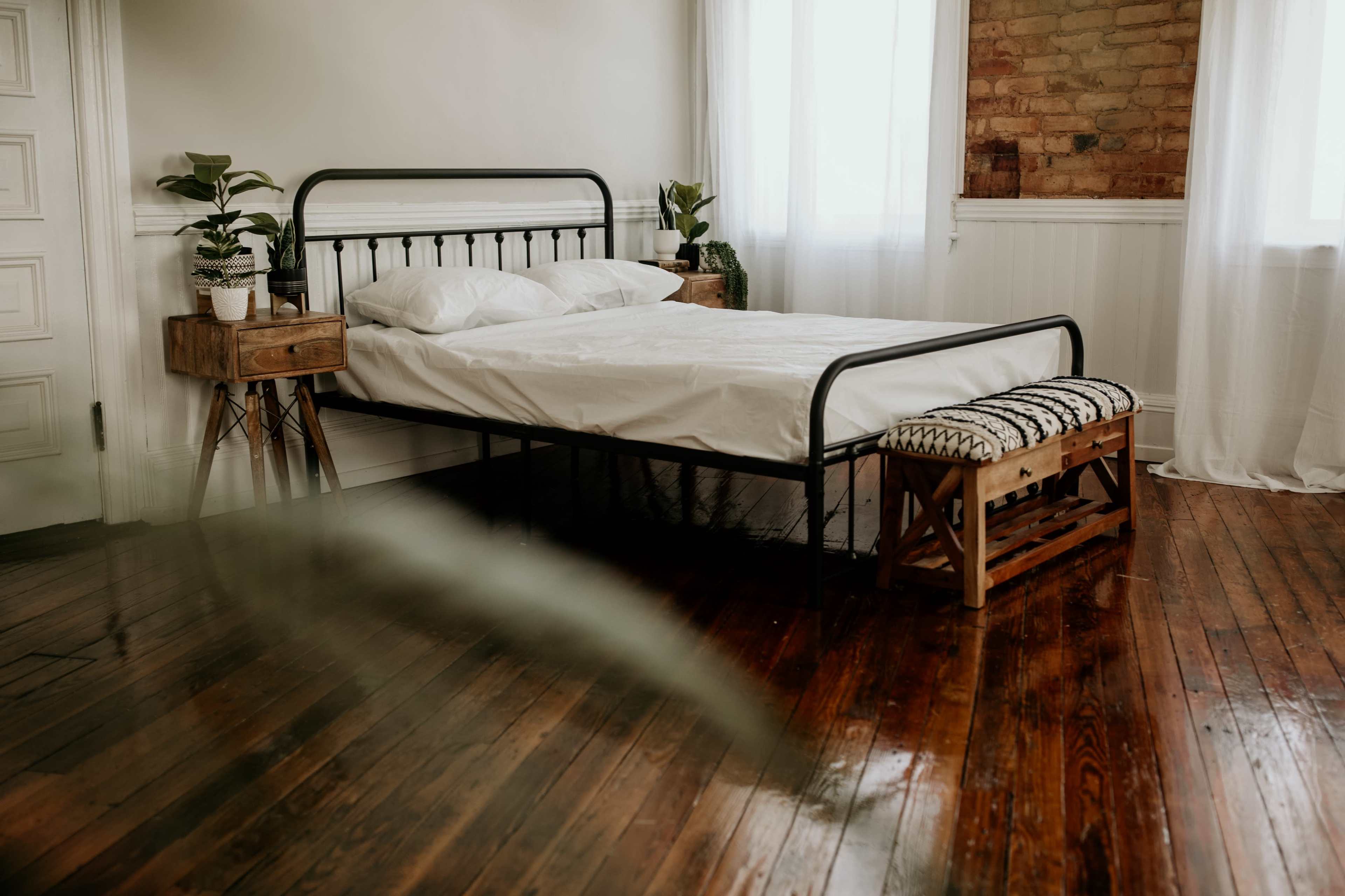 A minimalist bedroom features a metal bed frame with white bedding, wooden nightstands, and potted plants in a room with light-colored walls and wooden flooring.