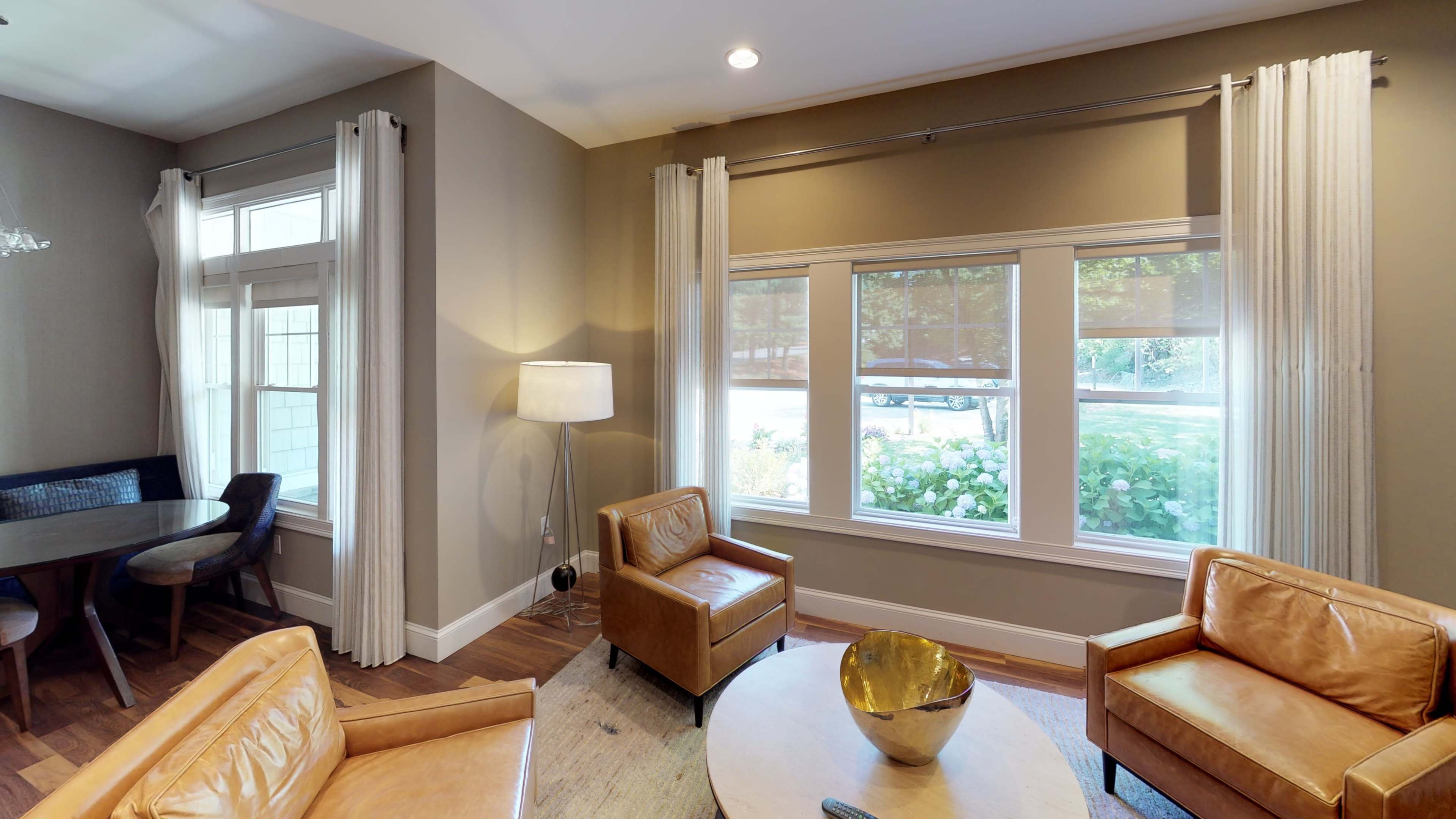 A living room with two brown leather chairs, a round table, a lamp, and large windows that allow natural light to enter.
