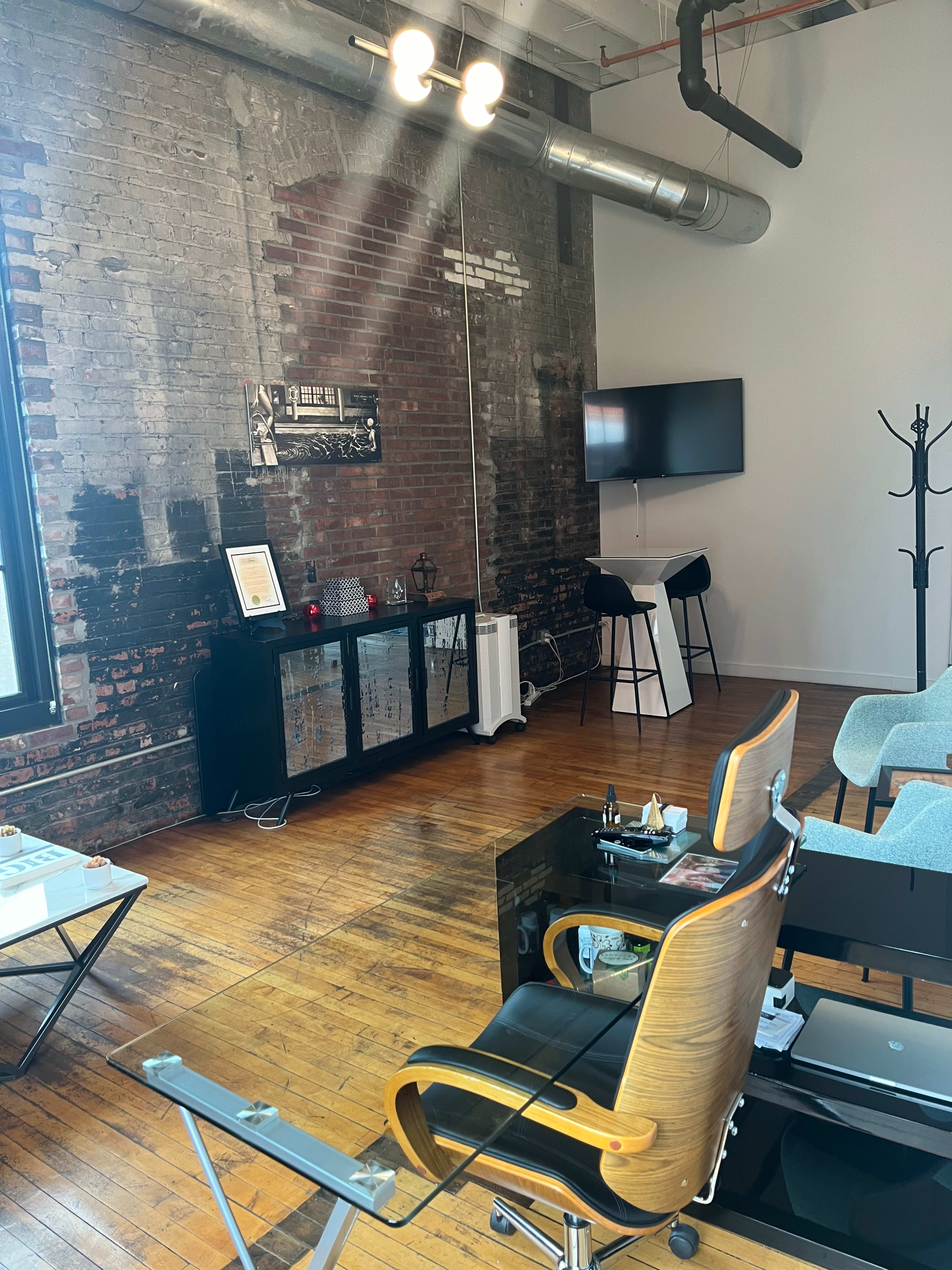 A modern office space with exposed brick walls, a television mounted on the wall, a wooden reception desk, and several chairs arranged for seating.