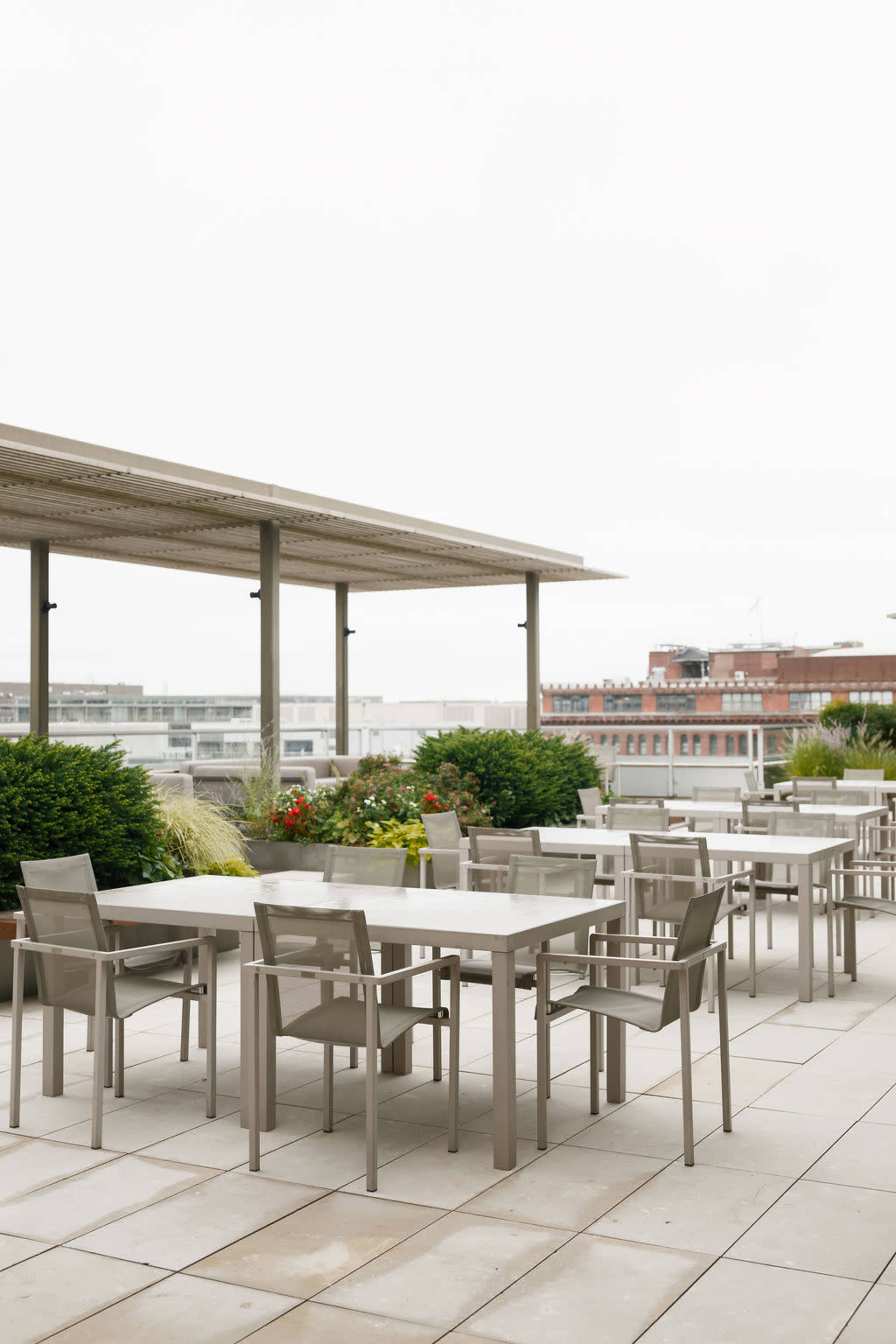 The image shows a spacious outdoor terrace featuring multiple rectangular tables and chairs surrounded by greenery and planters.