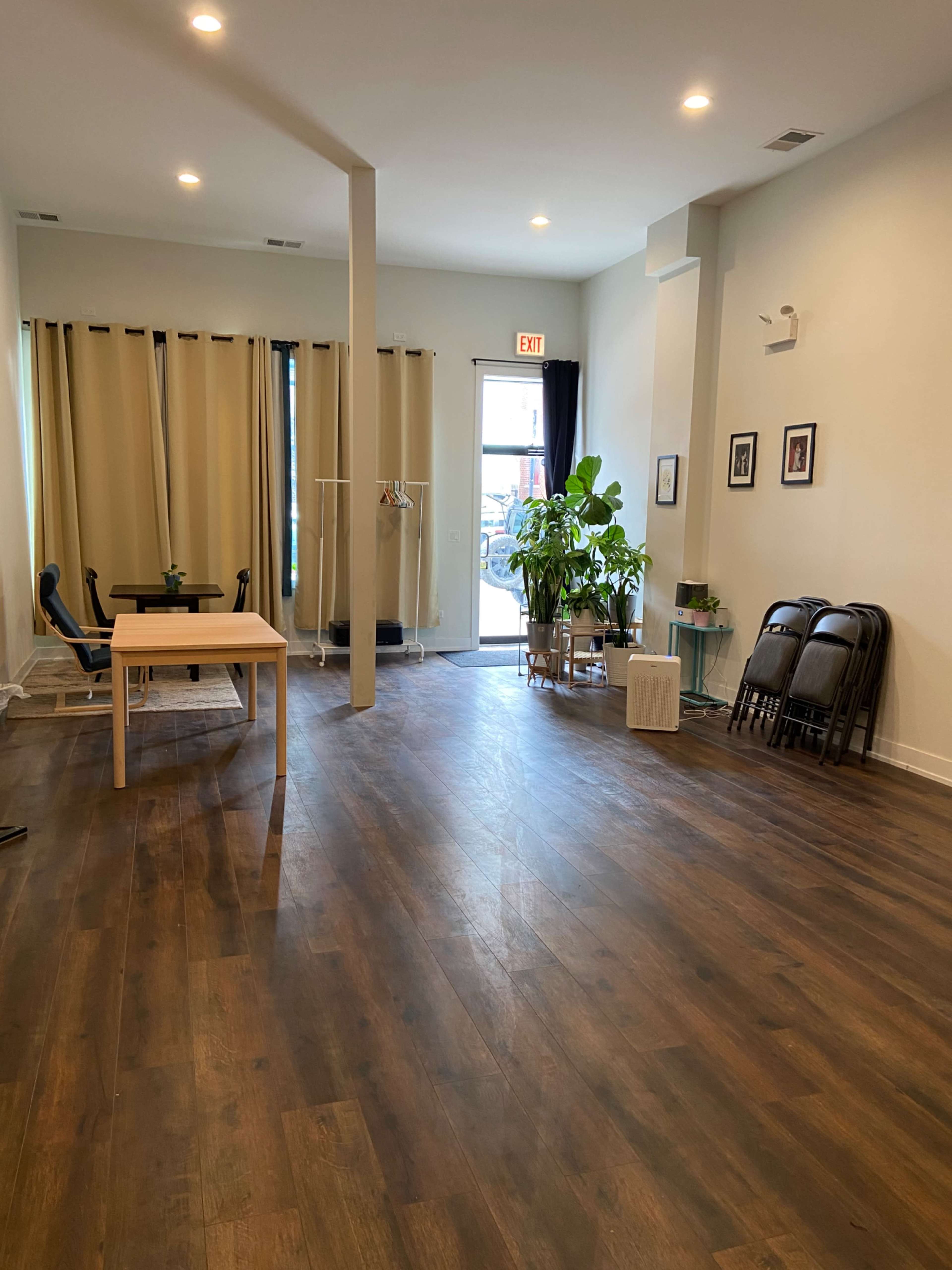 A spacious room with wooden flooring, a table, chairs, large windows with curtains, potted plants, and a door leading outside.