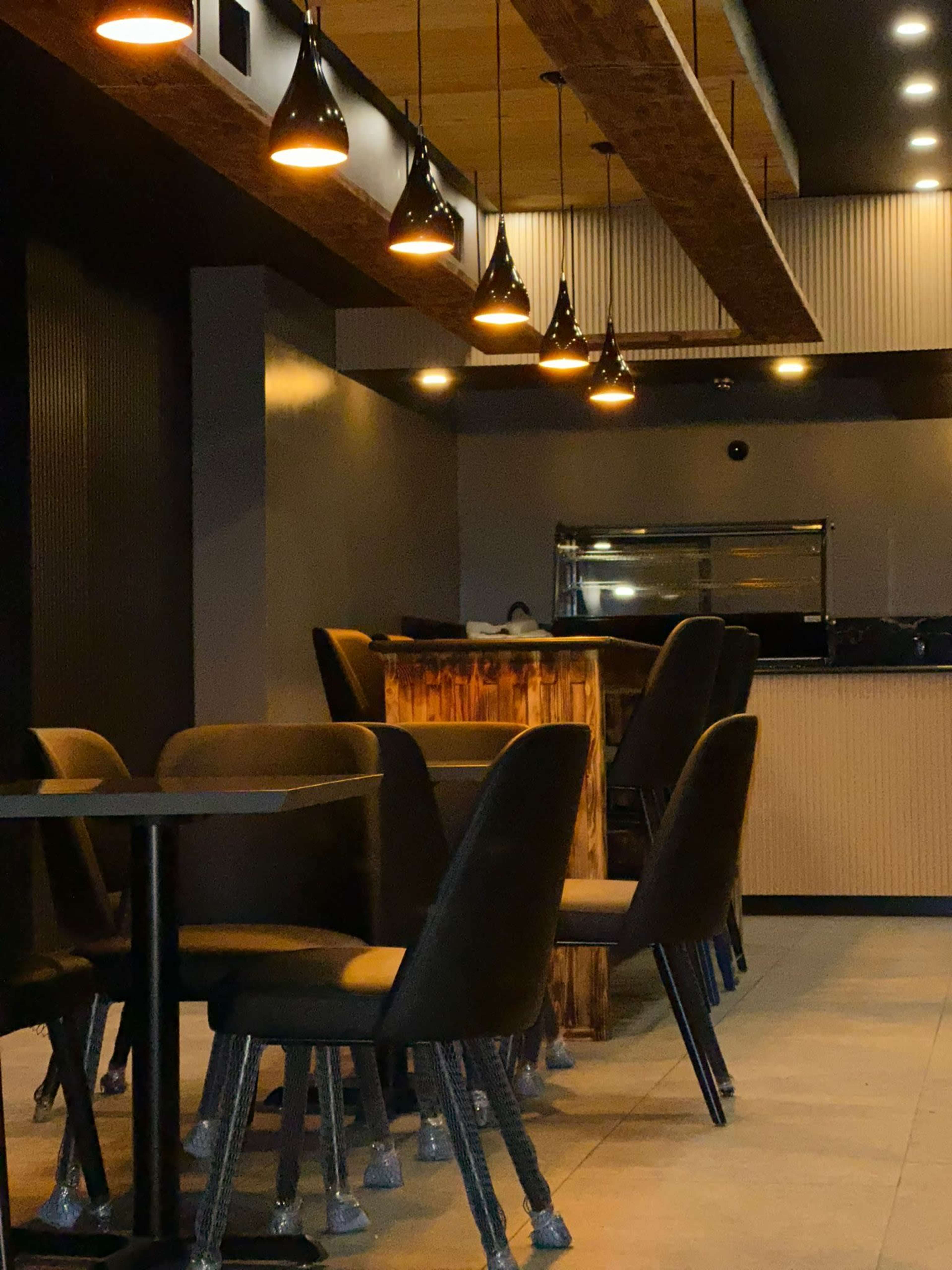 An interior of a restaurant with wooden elements, black chairs, and pendant lights hanging from the ceiling.