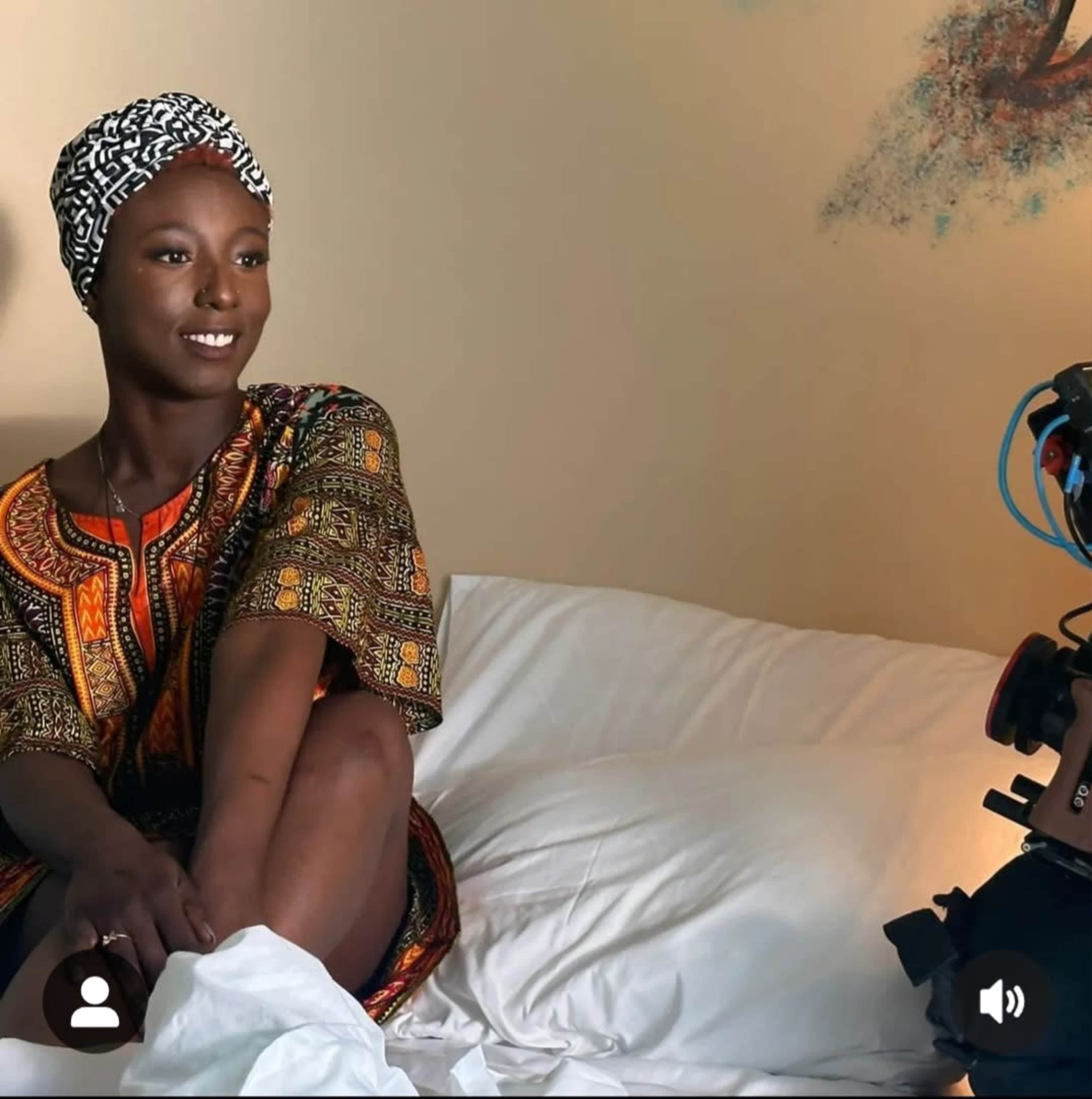 A woman in a patterned dress sits on a bed, looking at a camera set up beside her.