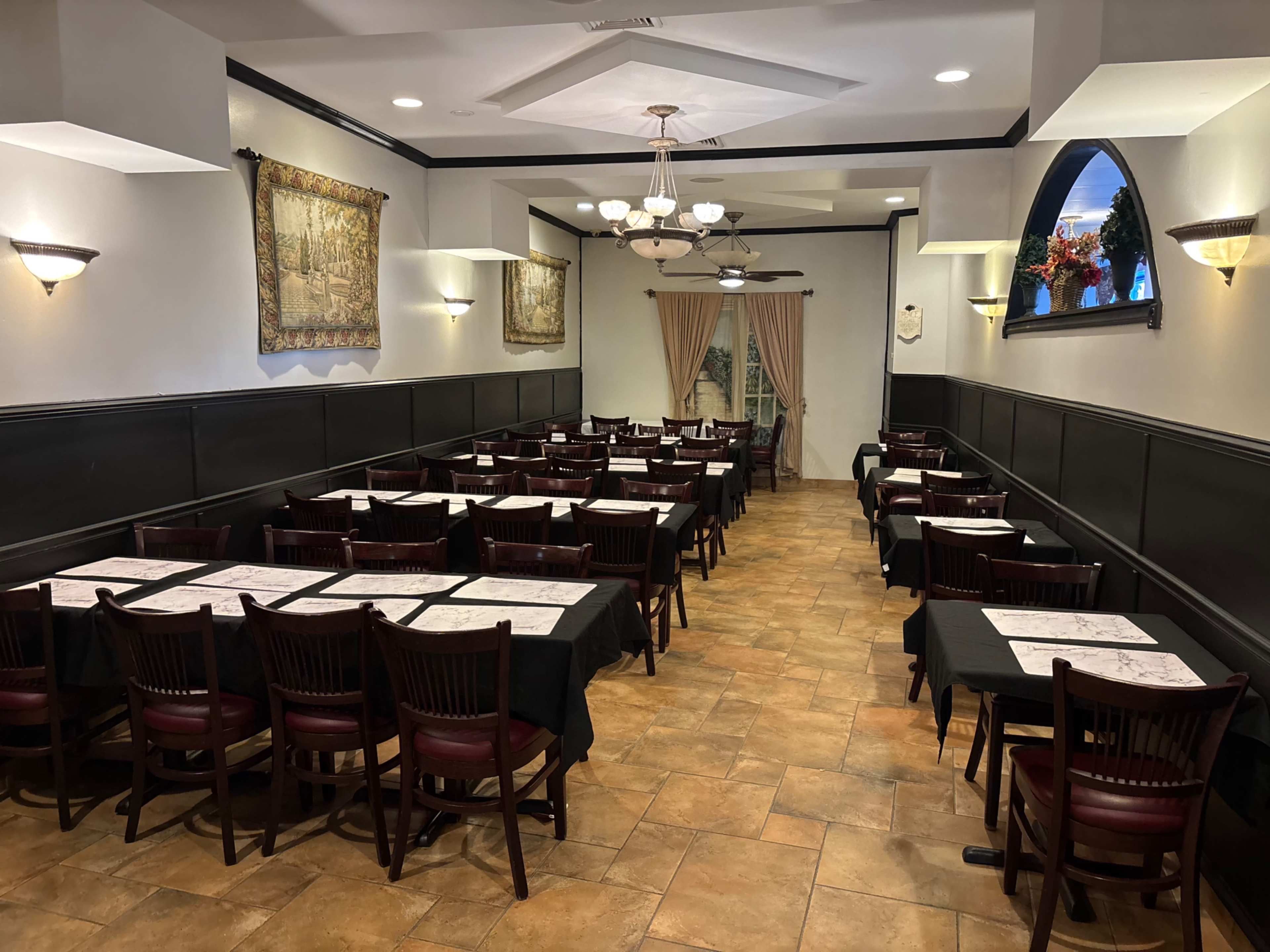 The image shows a spacious restaurant interior with several tables and chairs arranged in rows, featuring dark tablecloths and wall decorations.