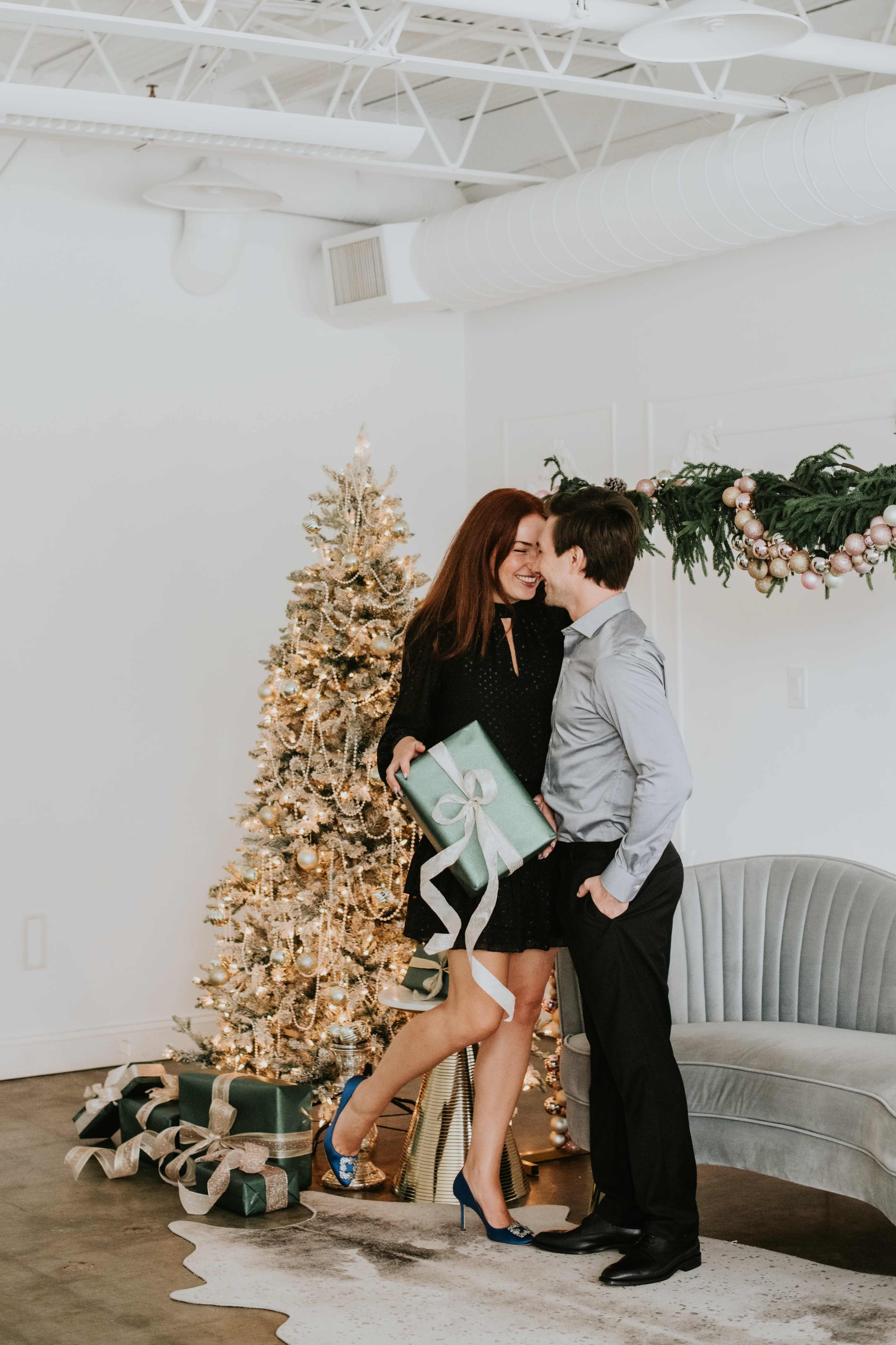 A couple exchanges a gift in front of a decorated Christmas tree in a festive indoor setting.