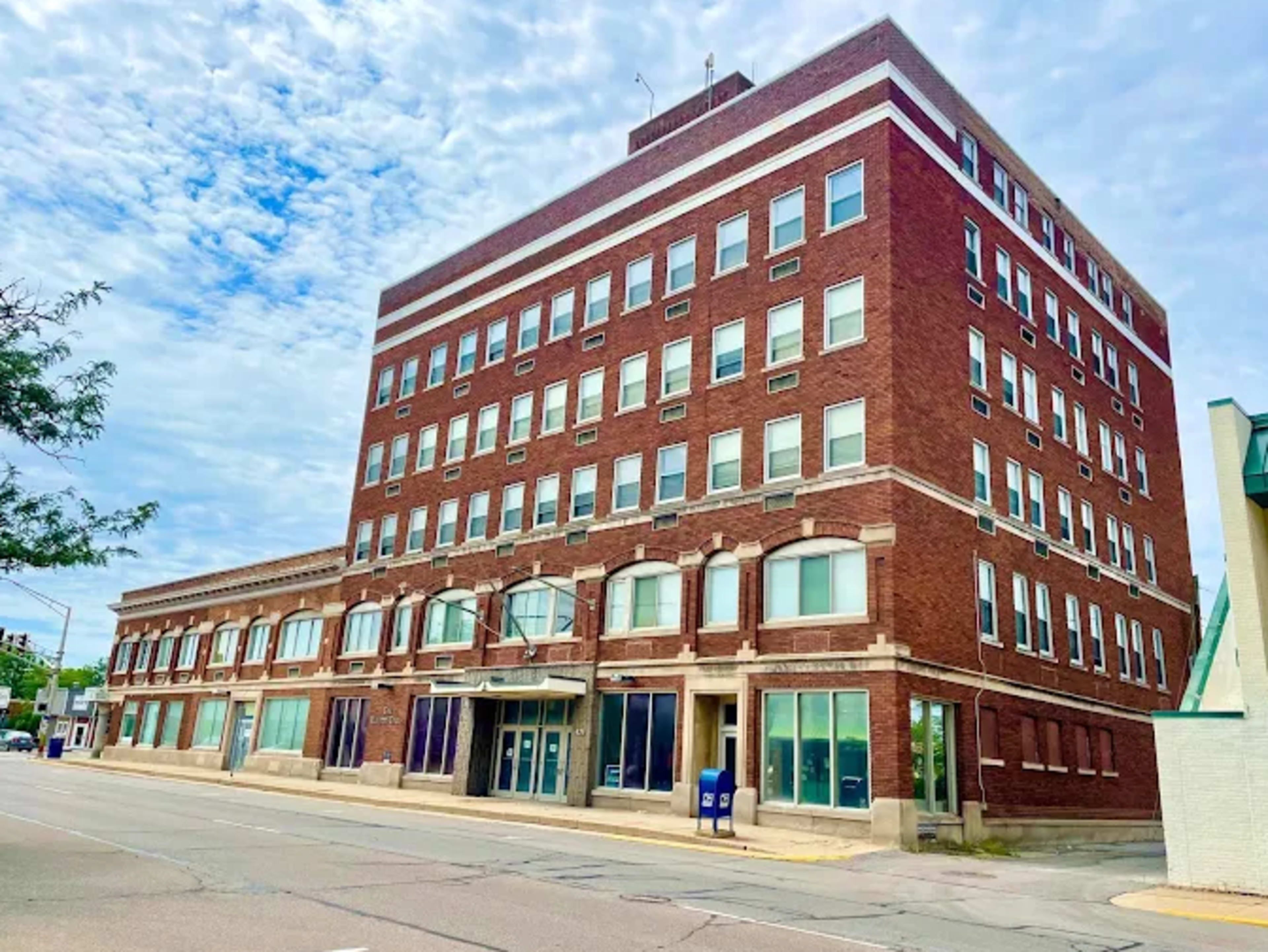 The image shows a four-story red brick building with multiple windows and a storefront entry facing a street.