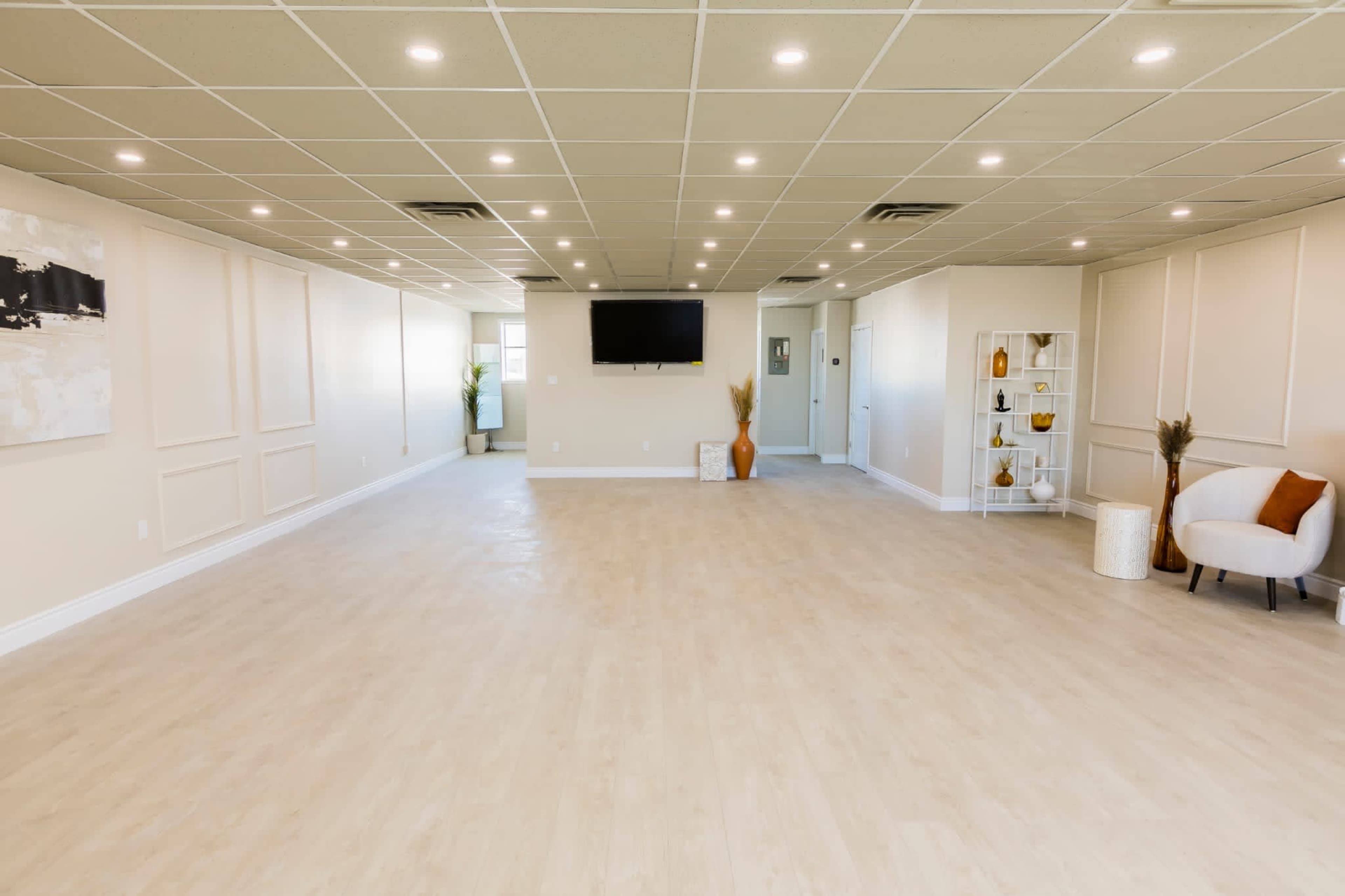 A spacious, well-lit interior room with modern decor, including a television on the wall and furnishings like a chair and decorative shelves.