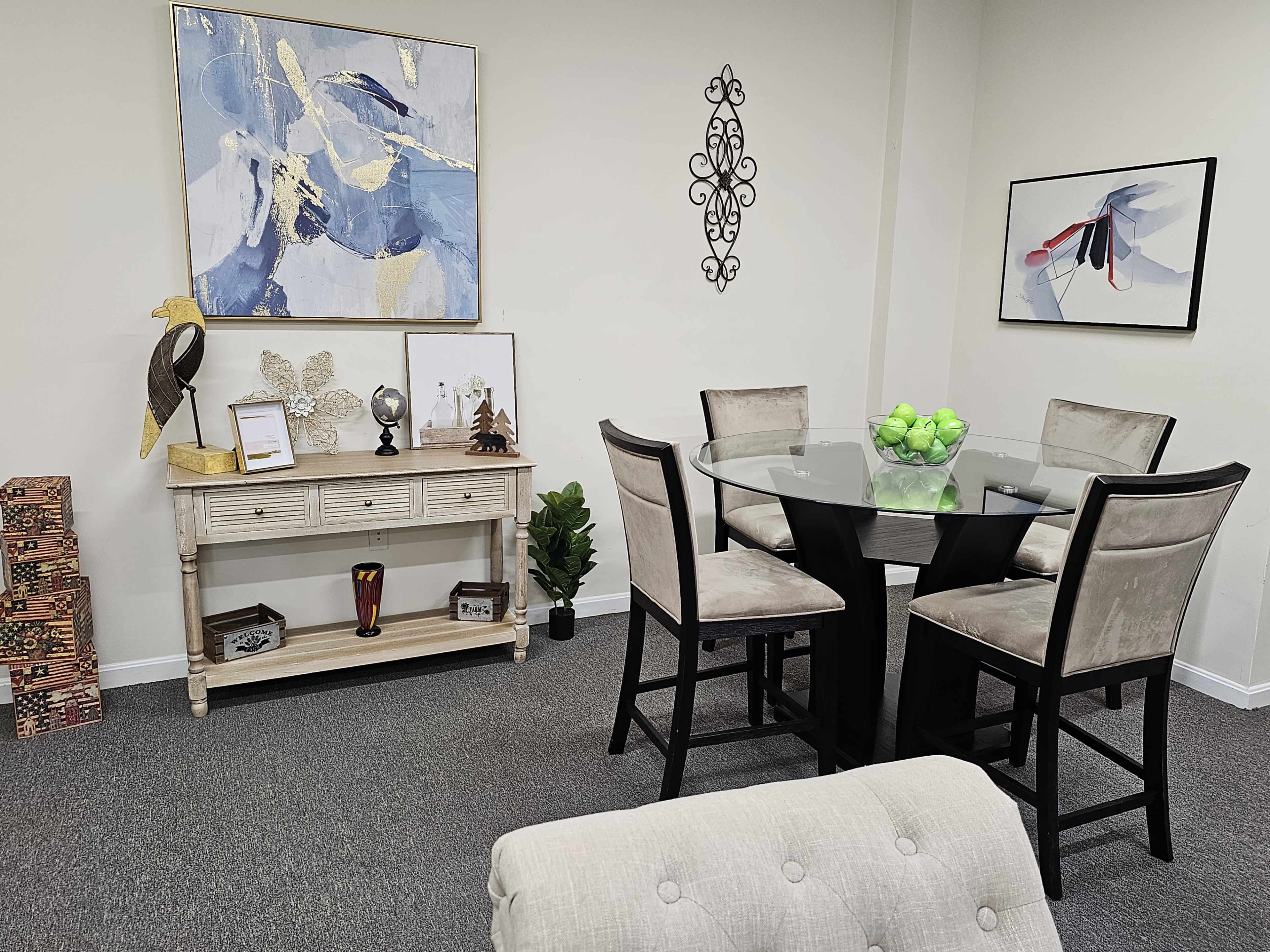 The image shows a neatly arranged modern dining area featuring a circular glass table surrounded by high-backed chairs, a side console table displaying decorative items, and artwork on the walls.