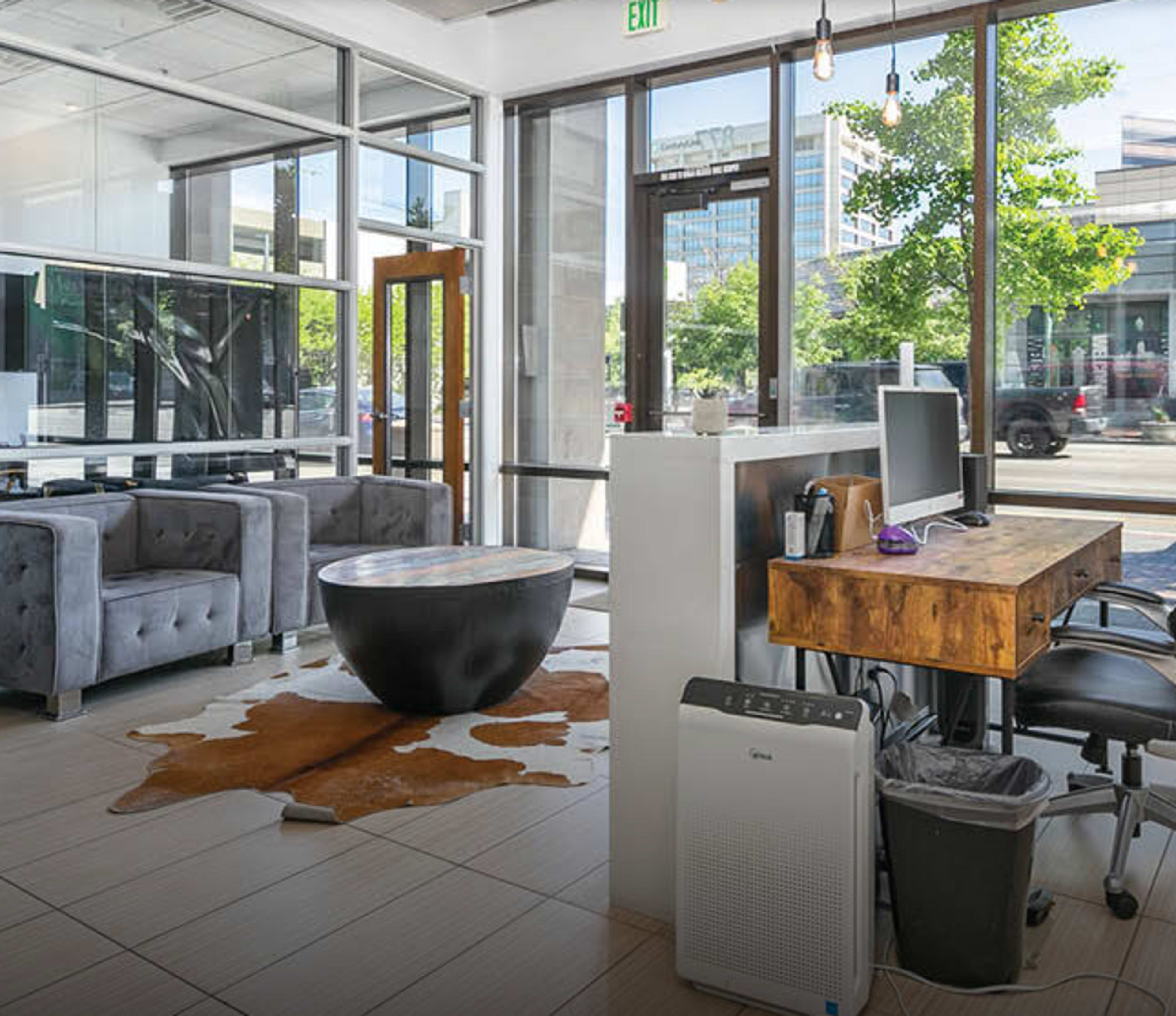 The image shows a modern office reception area featuring gray sofas, a circular black coffee table, and a workstation with a computer against large windows.