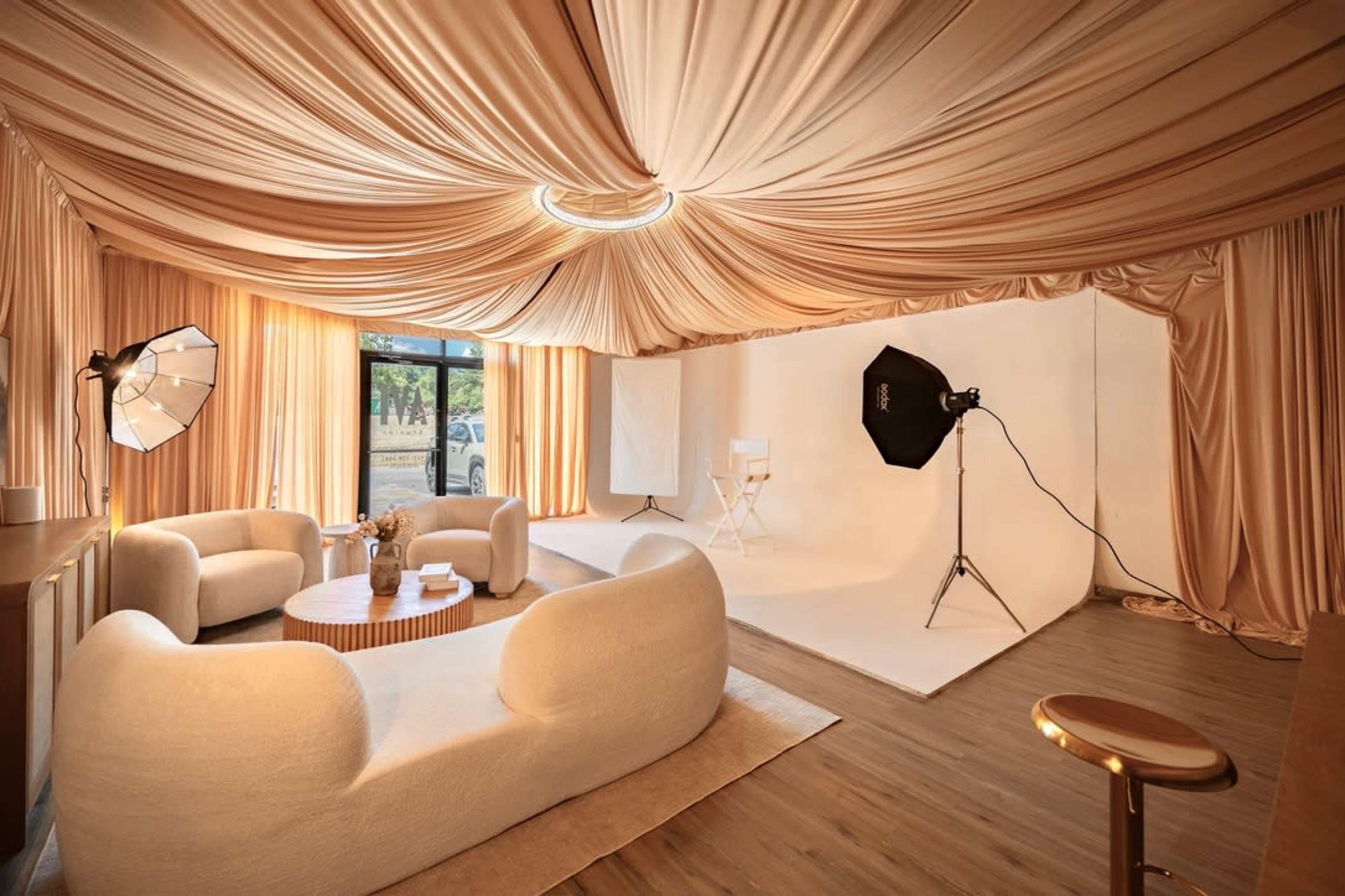 The image shows a spacious room with soft beige drapery on the ceiling and walls, featuring a seating area with rounded sofas and a photography setup with lights and a backdrop.