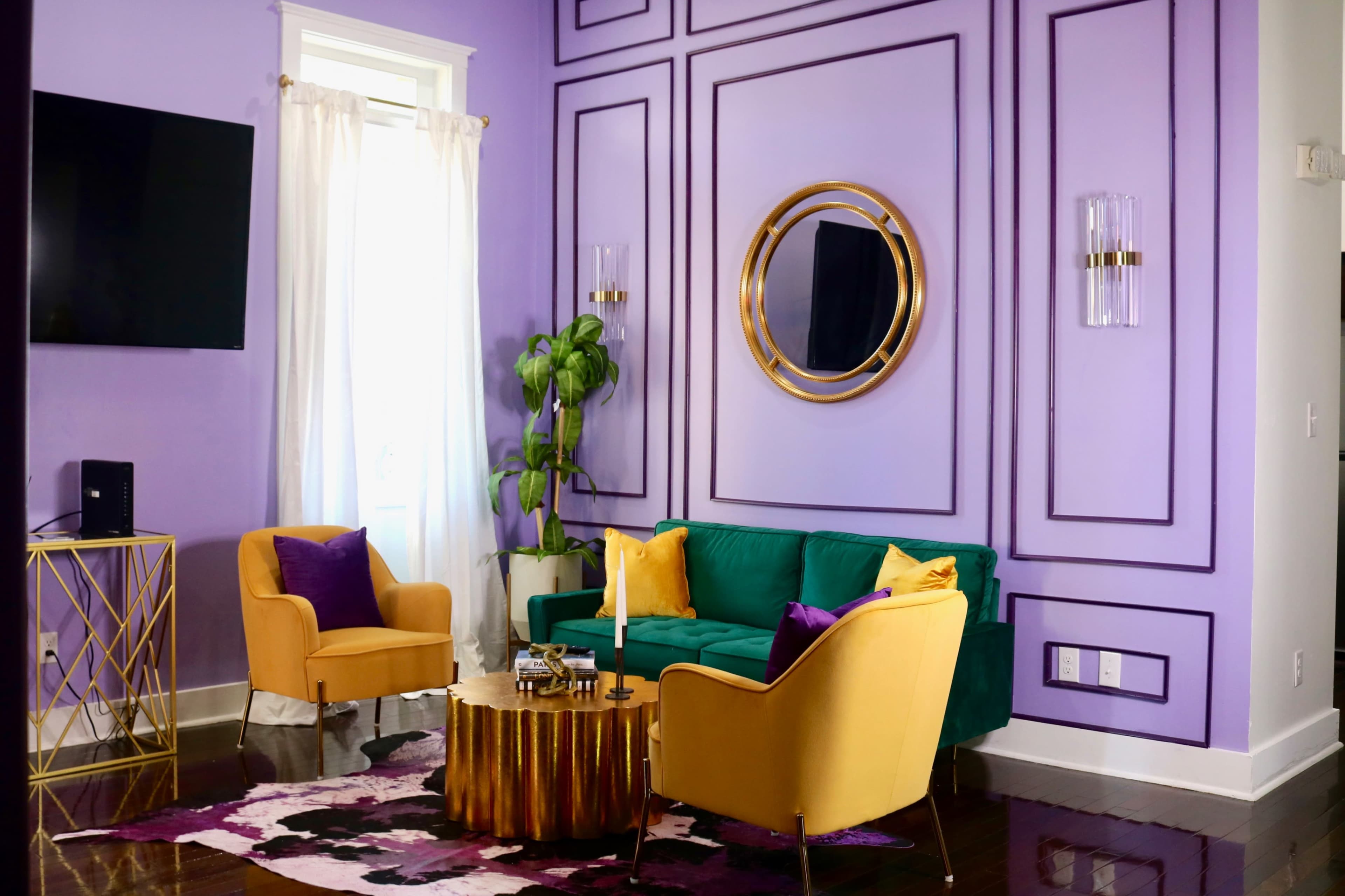 A brightly colored living room features a green sofa, yellow chairs, a round wooden coffee table, and purple walls with decorative molding.