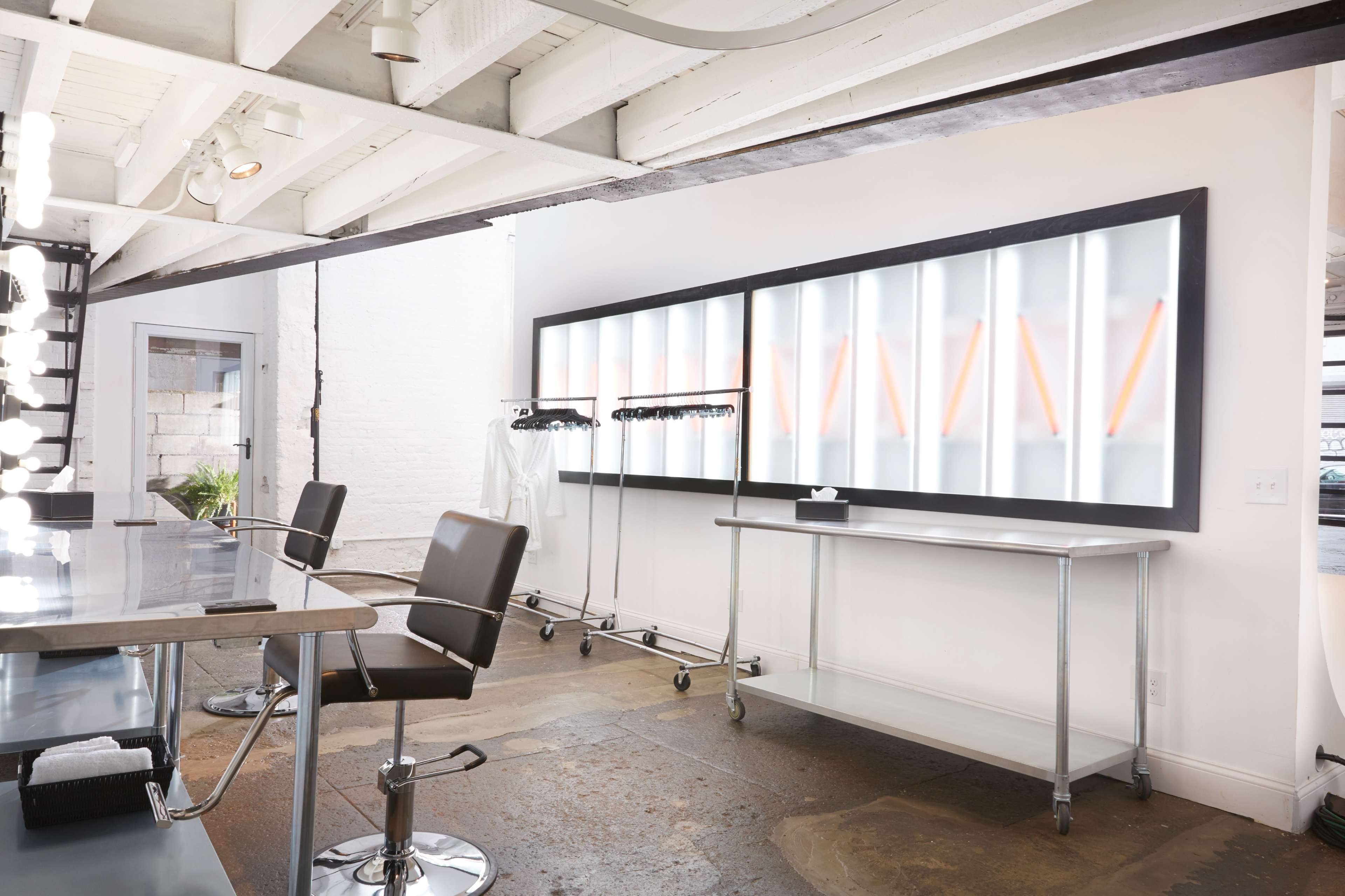 The image shows a modern salon interior featuring a large illuminated mirror, two black salon chairs, and a clothing rack against a white wall.