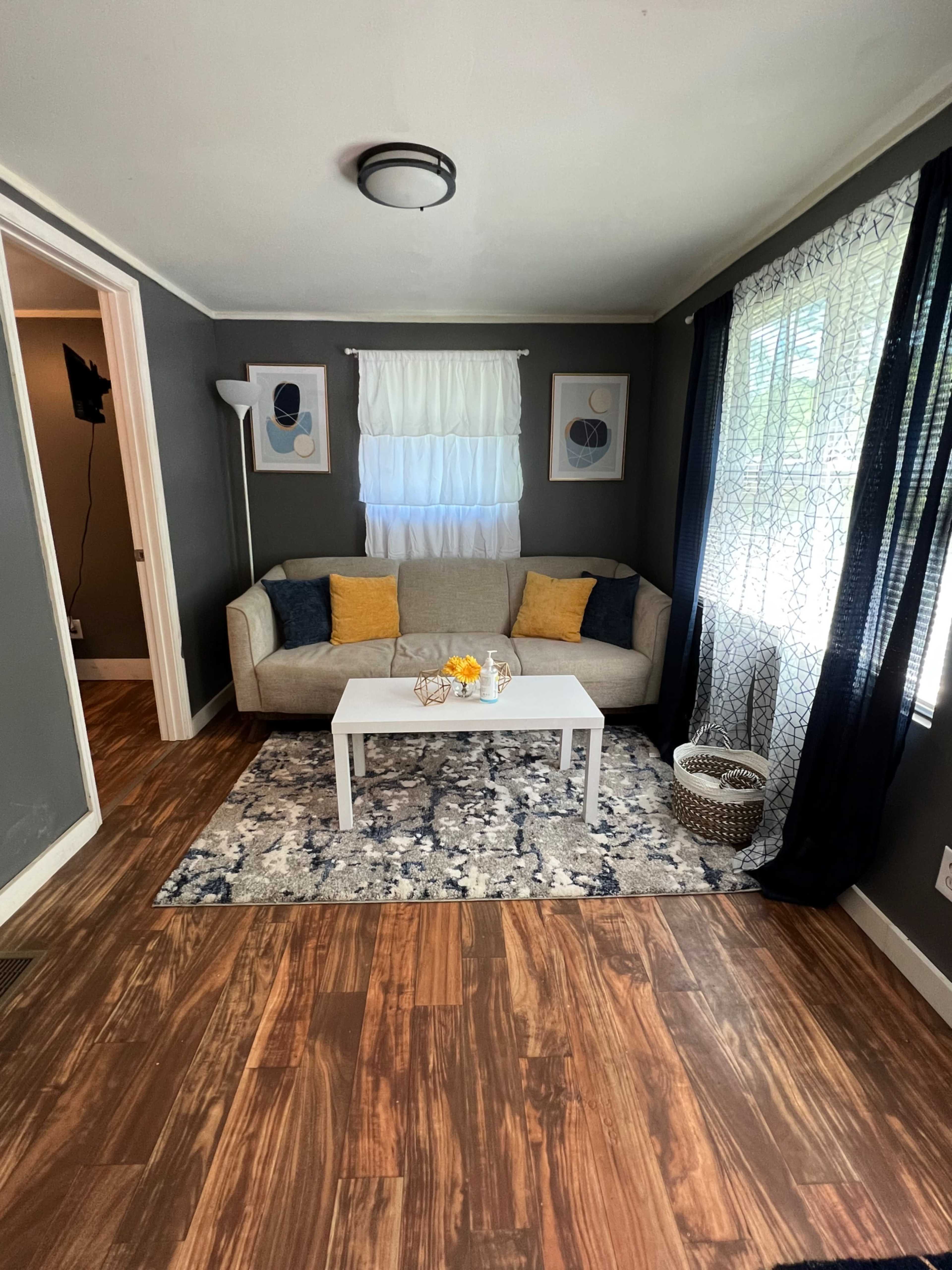 A cozy living room featuring a light gray sofa, a white coffee table, and a patterned area rug, with large windows draped in sheer and dark curtains.