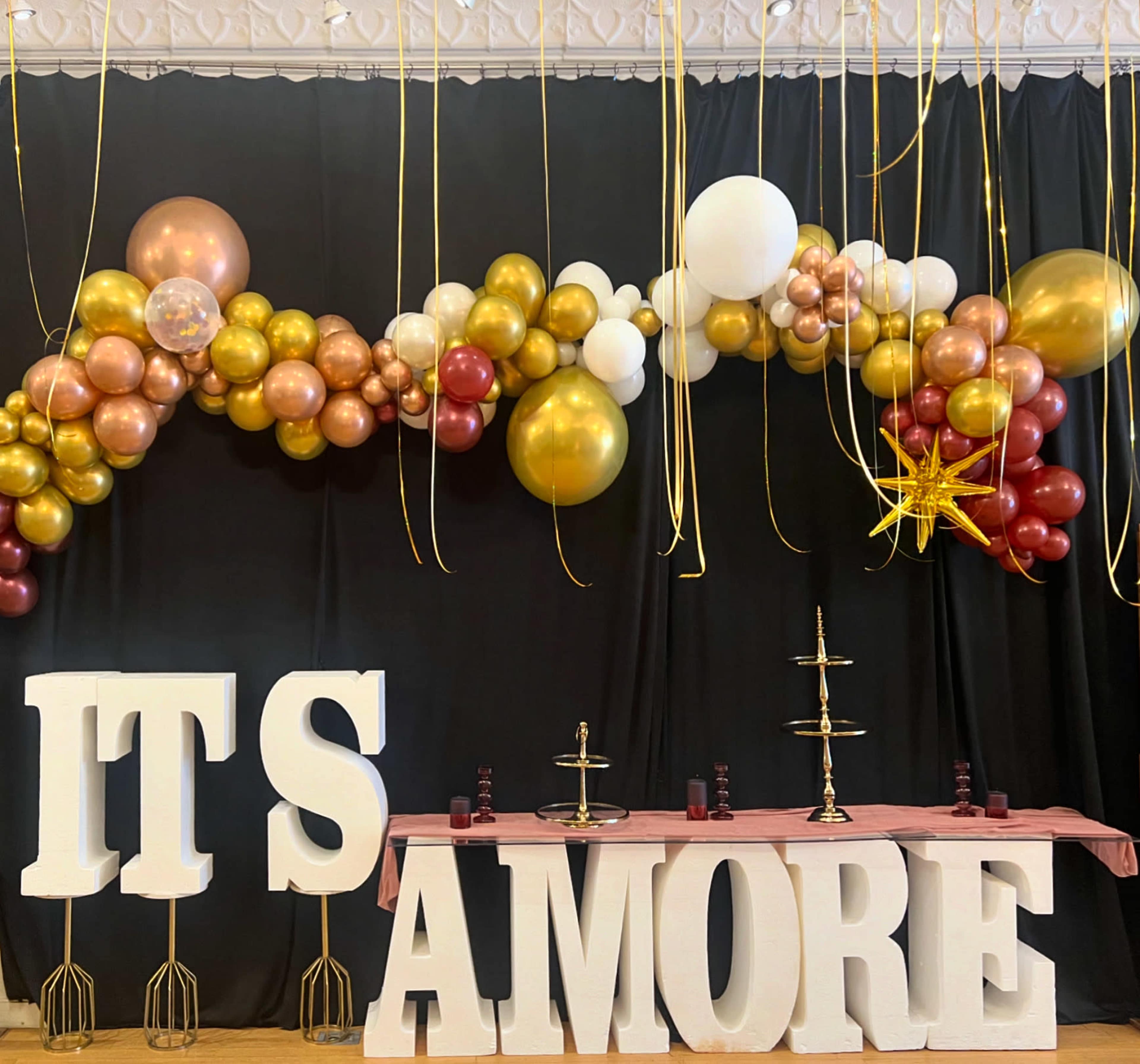 The image features a festive display with a large balloon arch in gold, white, and maroon, and oversized letters spelling "ITS AMORE" on a table with decorative stands beneath a black backdrop.