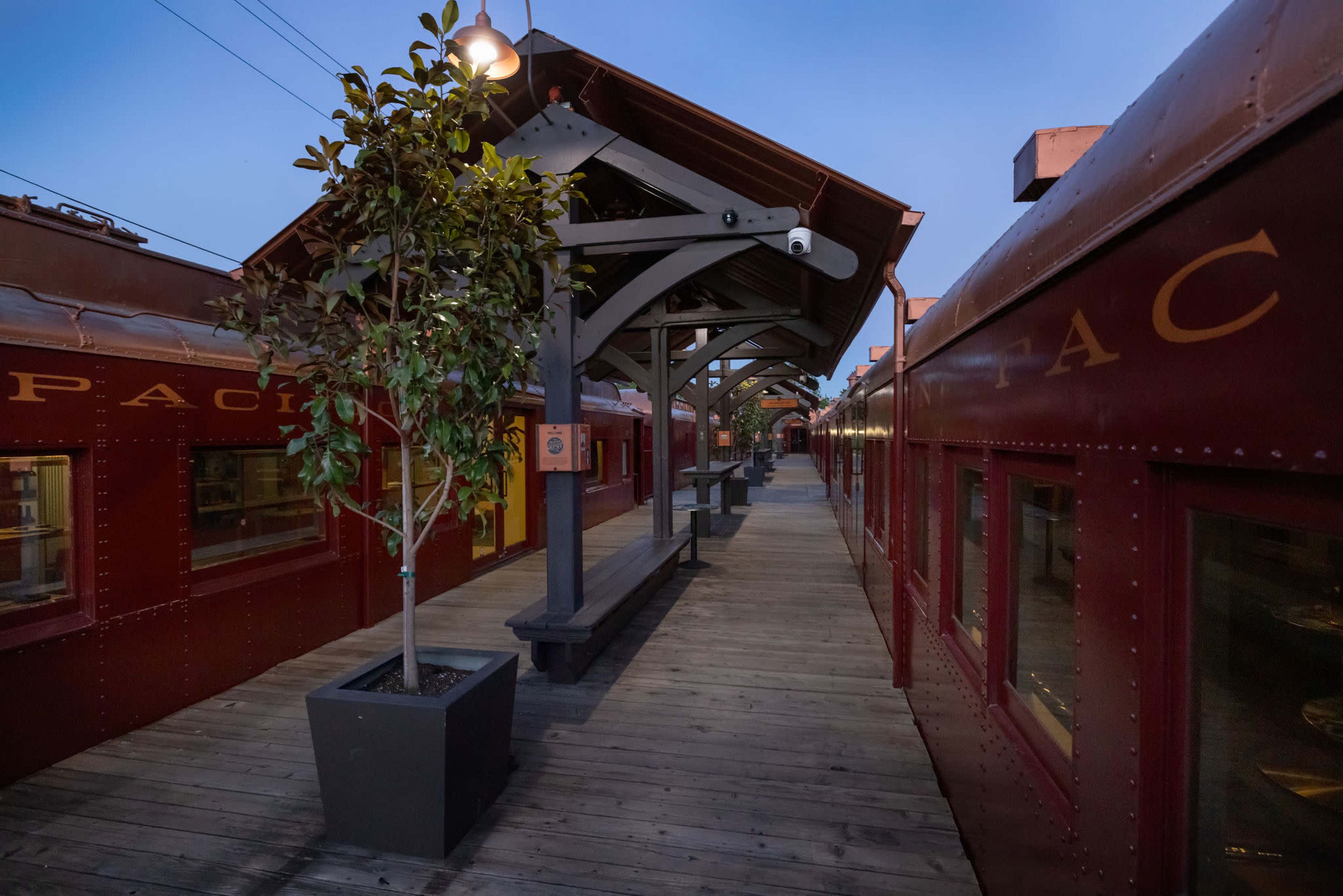 The image shows a wooden walkway between two vintage red train cars, with a planter and overhead sheltering structure in a dimly lit environment.